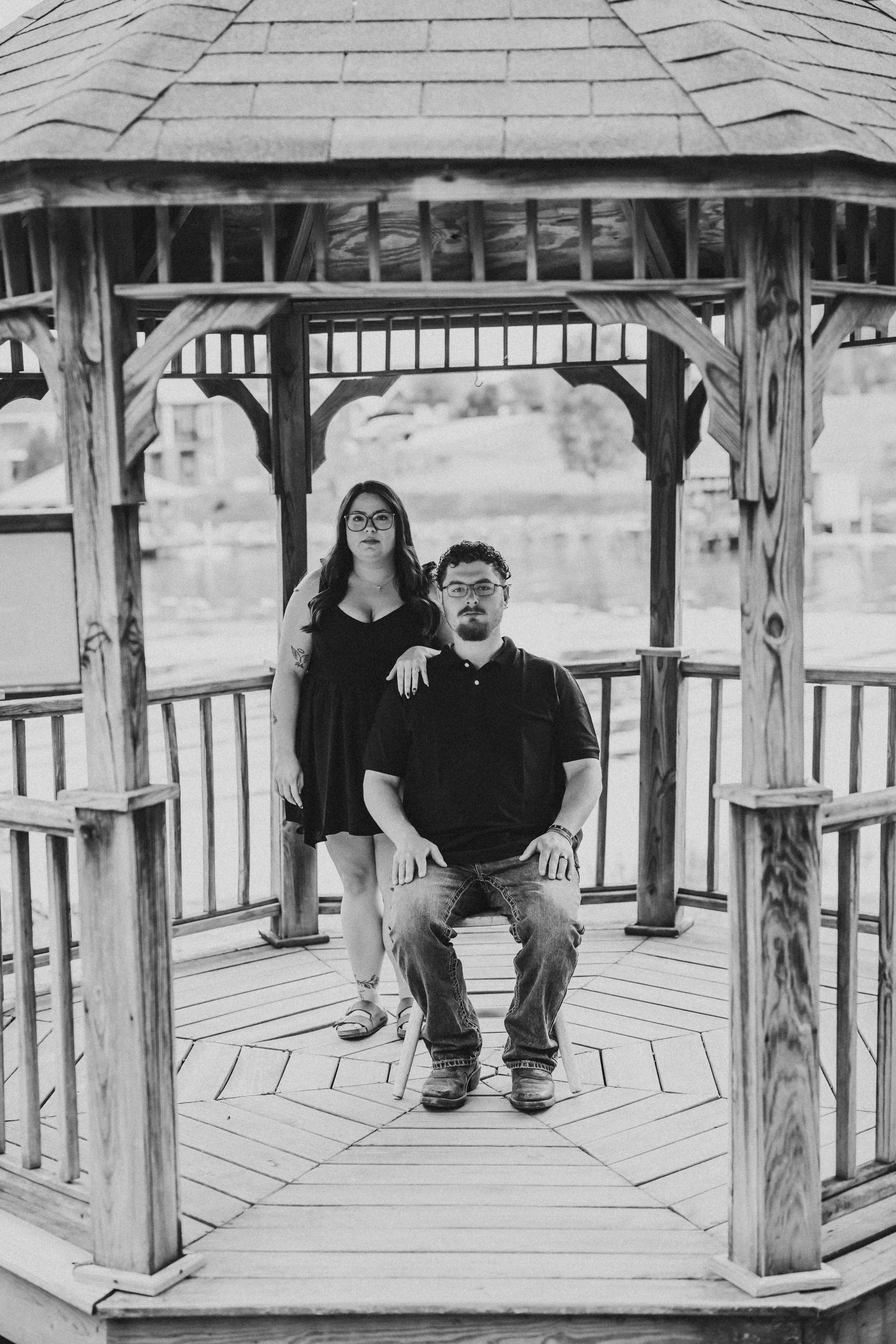 Two people, a woman and a man, standing and sitting on a wooden gazebo. The woman has long dark hair, glasses, and a black dress. The man has curly hair, glasses, a black shirt, and jeans. The gazebo has a shingled roof and decorative wooden supports