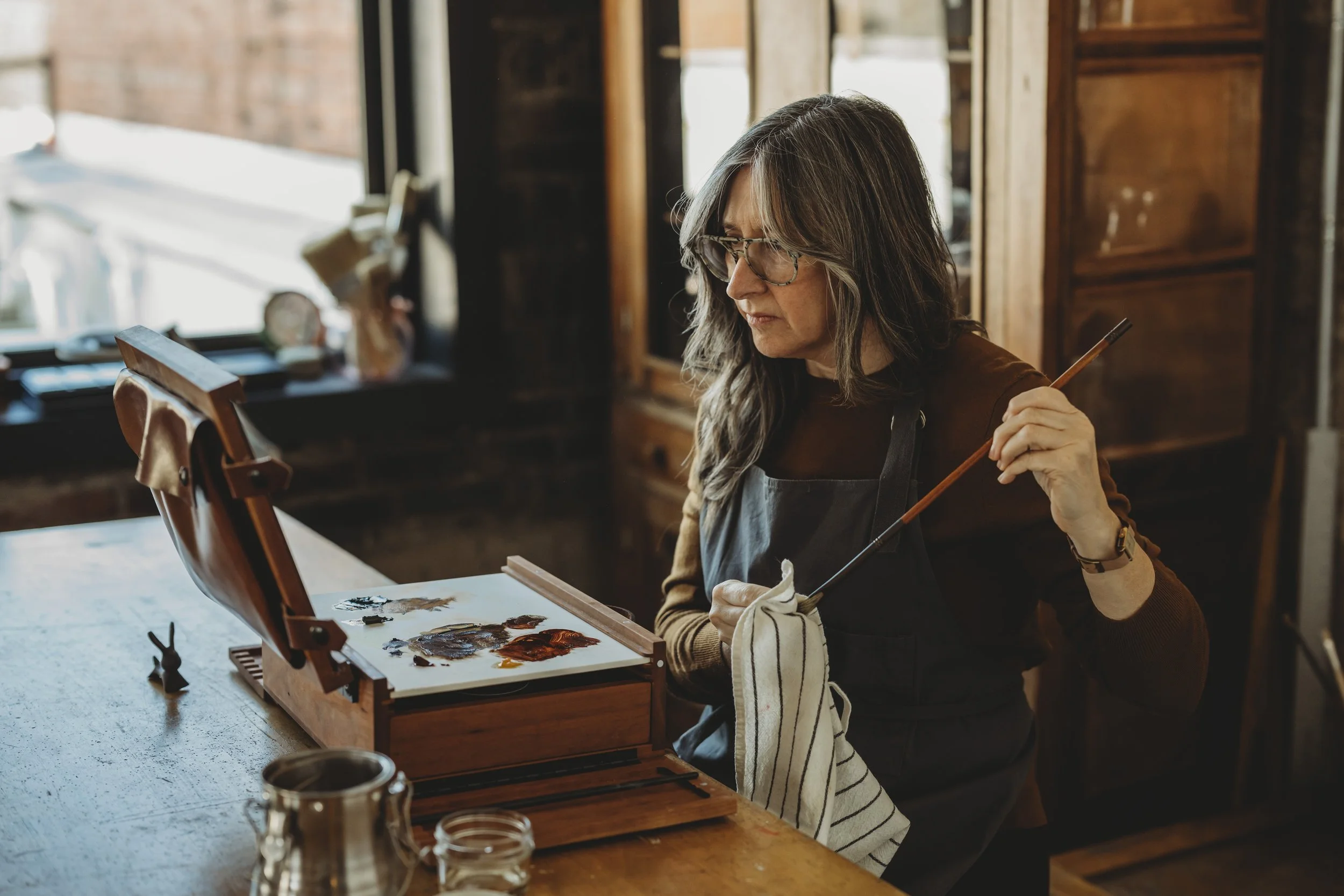 A woman wearing glasses and a dark apron, holding a paintbrush and a cloth, is working on a painting in a cozy, rustic studio with wooden furniture and large windows letting in natural light.