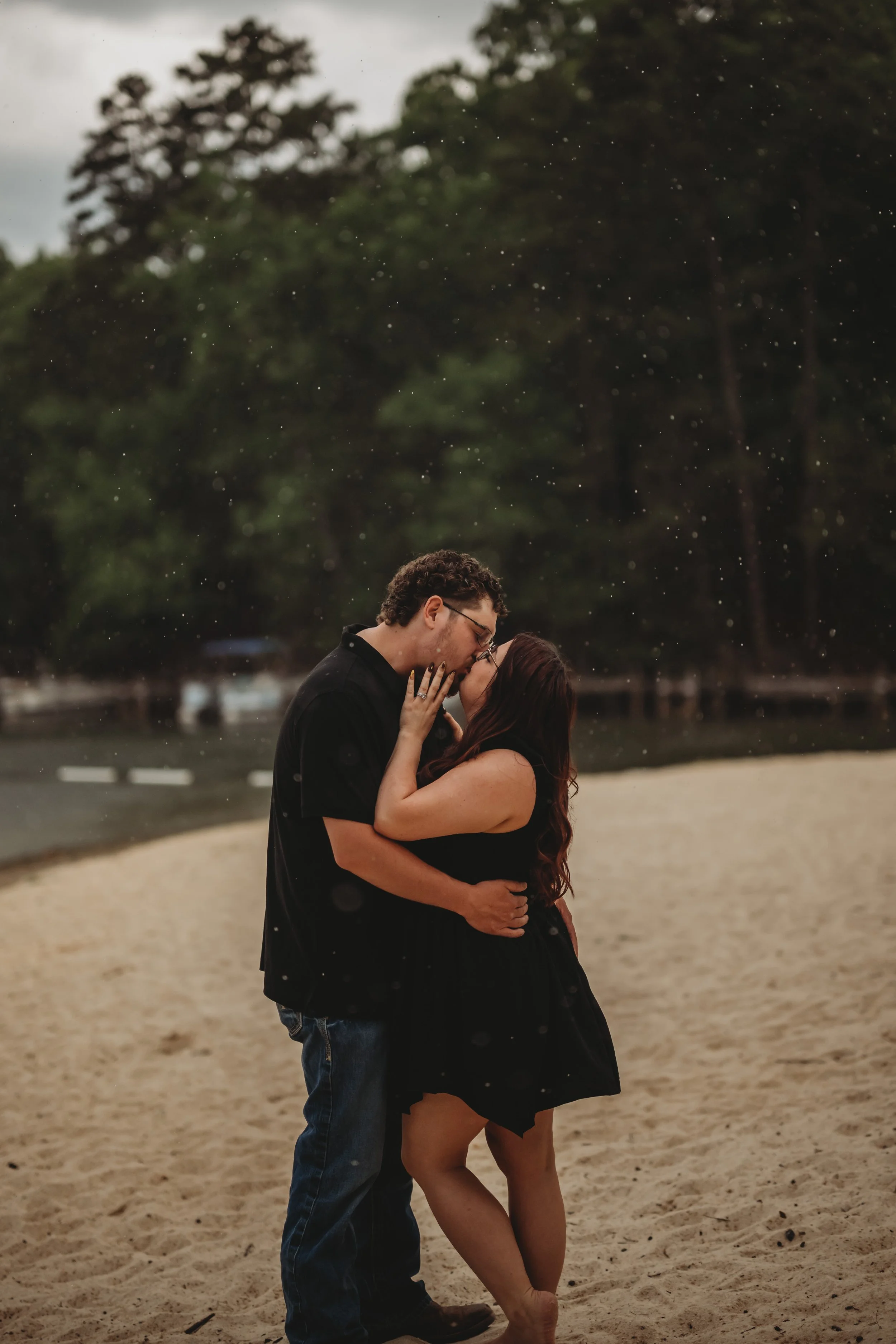 A couple sharing a kiss on a beach, with trees and water in the background, during overcast weather.