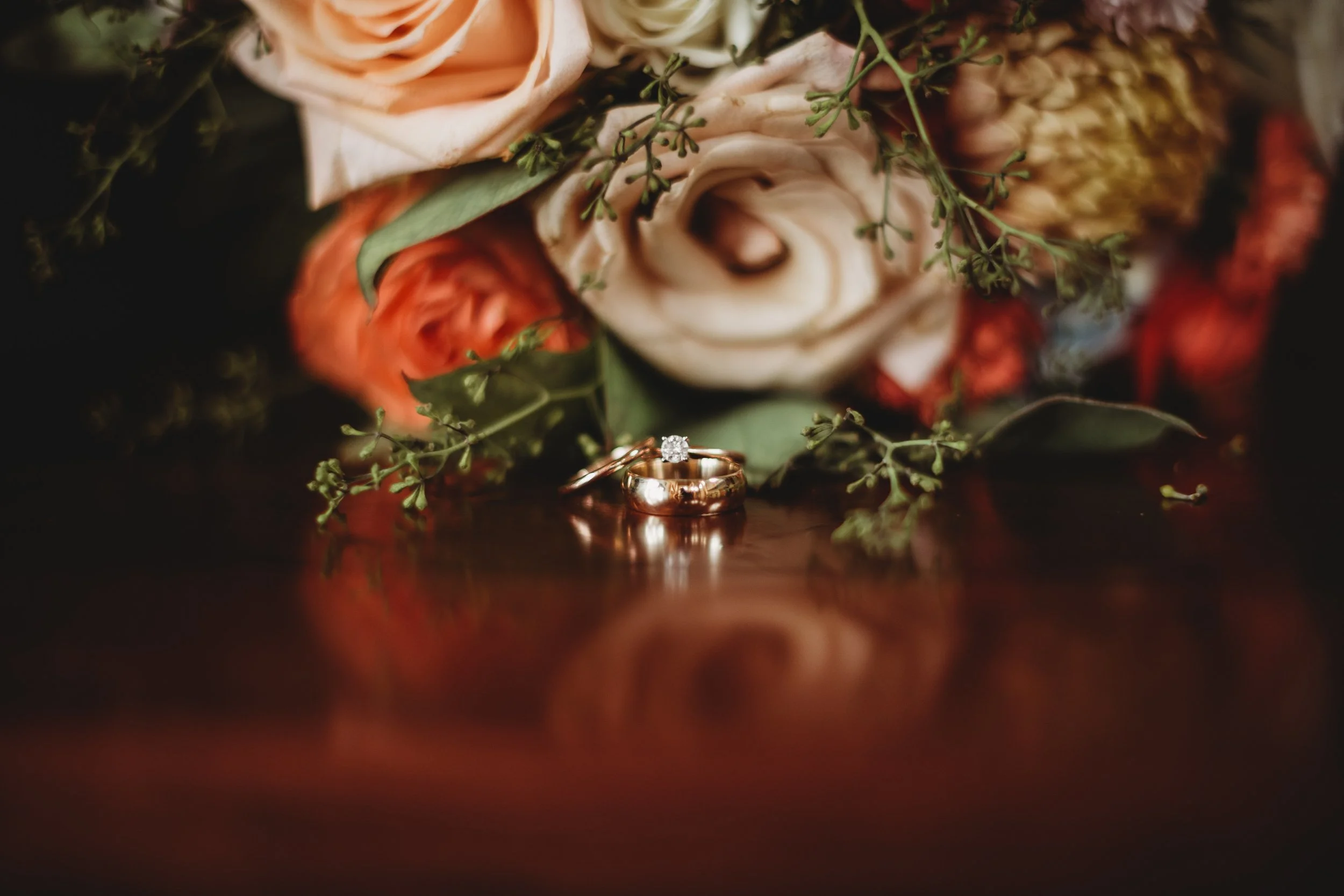 Close-up of a wedding ring and engagement ring on a reflective wooden surface with a soft floral bouquet in the background.