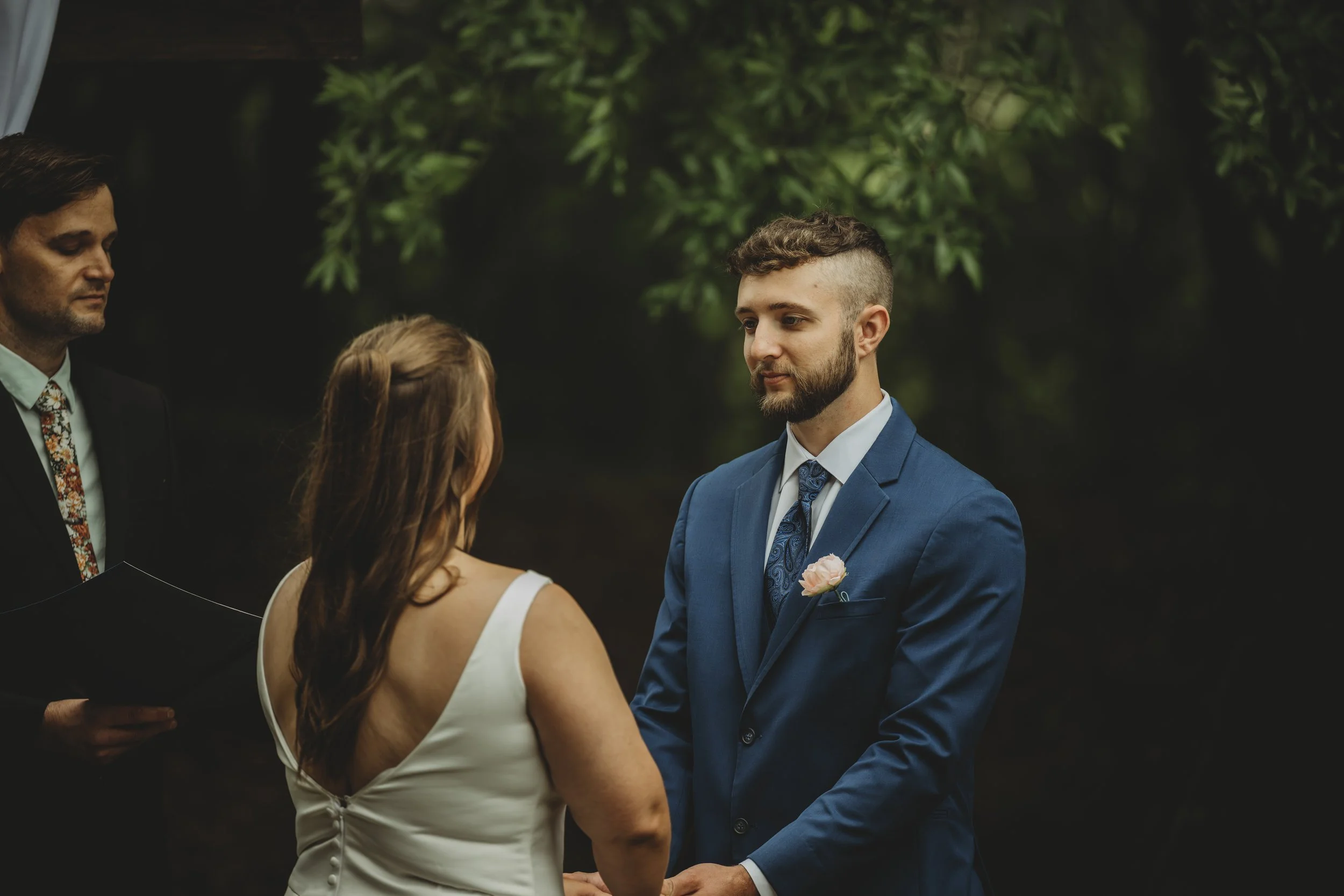 A man and woman exchanging vows at a wedding ceremony outdoors, with a man reading from a book in the background, surrounded by trees.