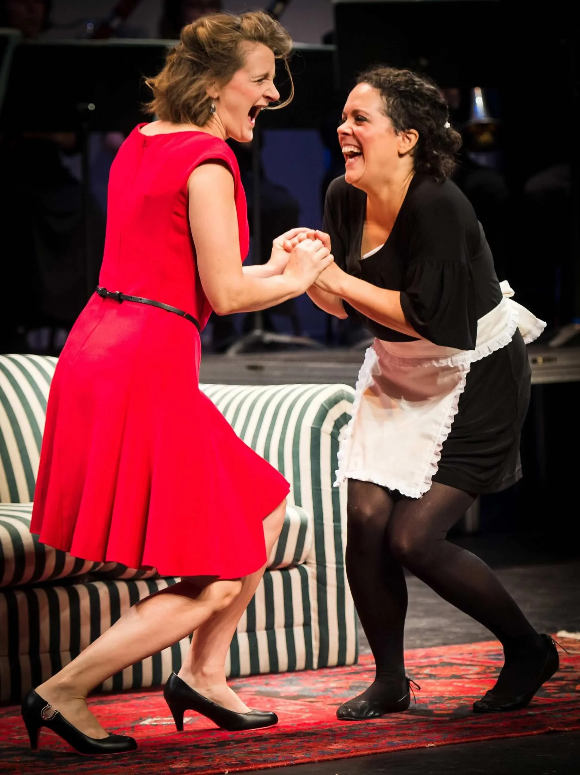 Deux femmes en costume scintillant, souriantes et le visage expressif, se tenant par les mains en riant, avec une personne tenant une perche filmant la scène, dans un décor intérieur avec un tapis rouge.