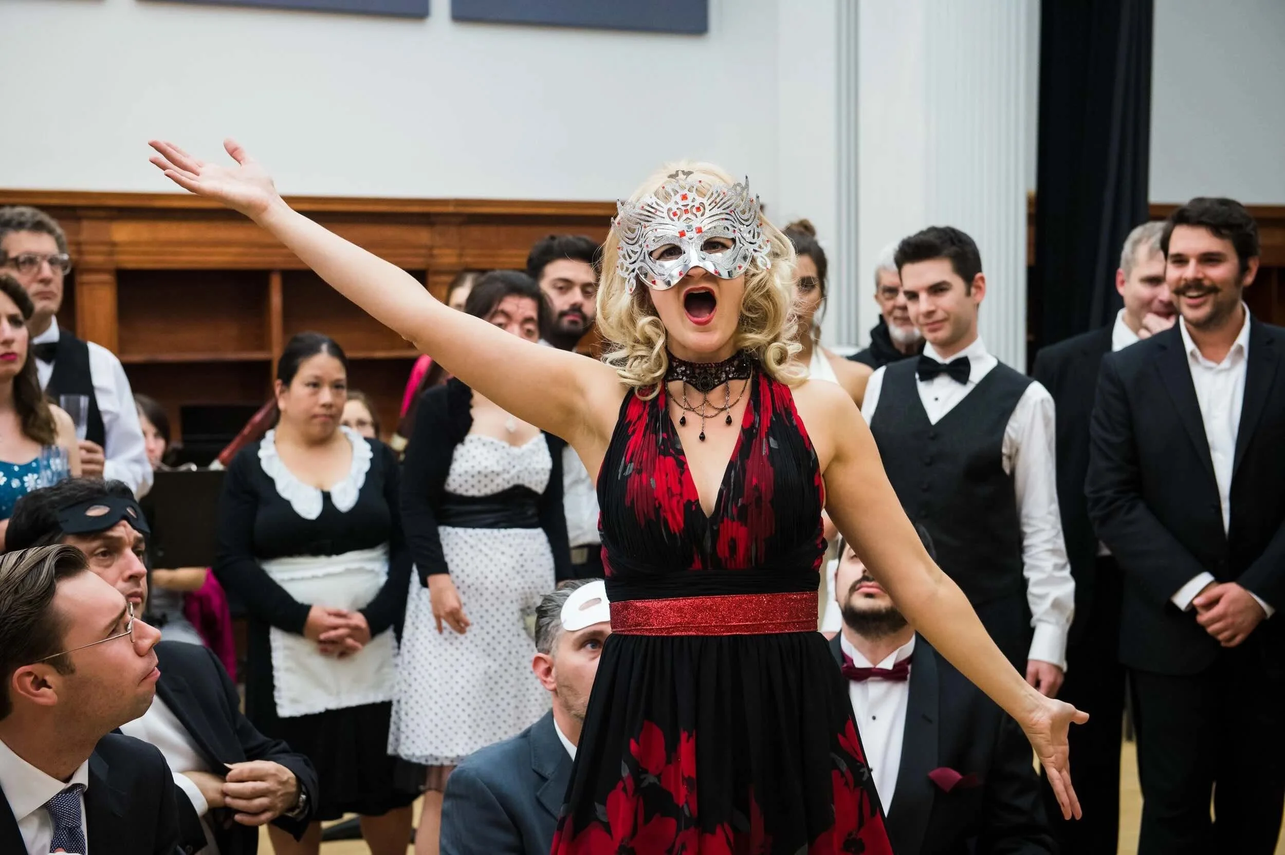 Sophie De Cruz portant un masque élégant et une robe rouge et noire, au centre d'une foule lors d'un événement social ou une fête, avec plusieurs personnes en costume traditionnel autour d'elle.