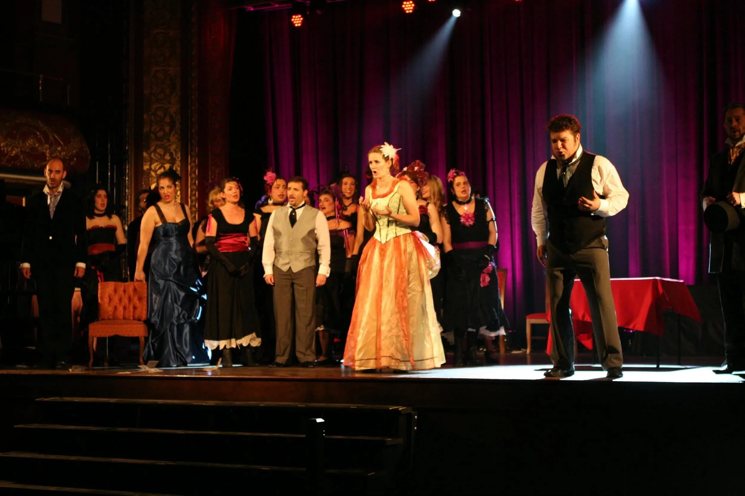 Acteurs sur scène d'une pièce de théâtre, certains chantent ou parlent, avec un décor de théâtre à fond violet et des éclairages lumineux.