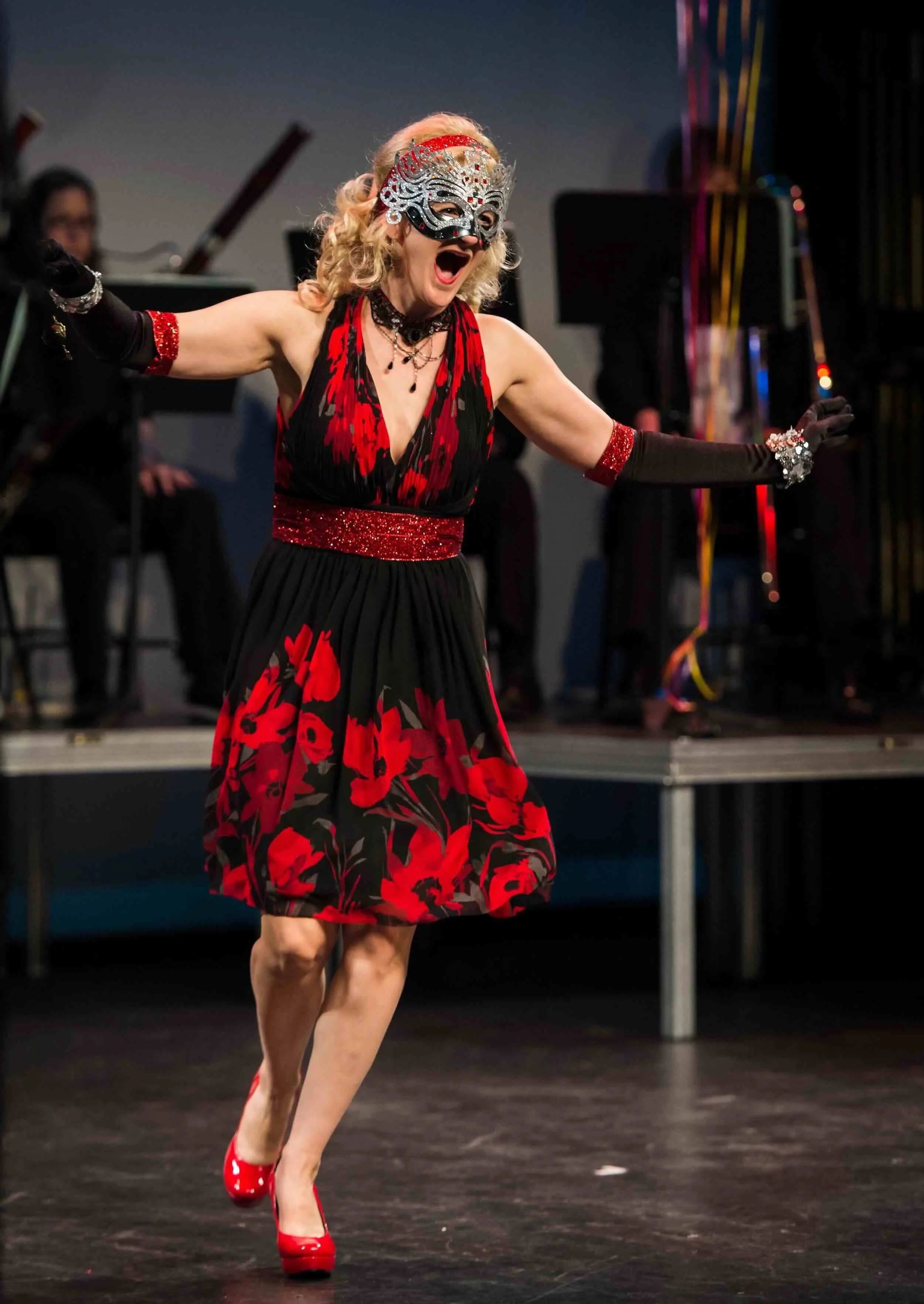 
Sophie De Cruz, portant un masque de carnaval, une robe noire à motifs rouges, des gants longs noirs et des chaussures à talons rouges, avec un orchestre en arrière-plan.