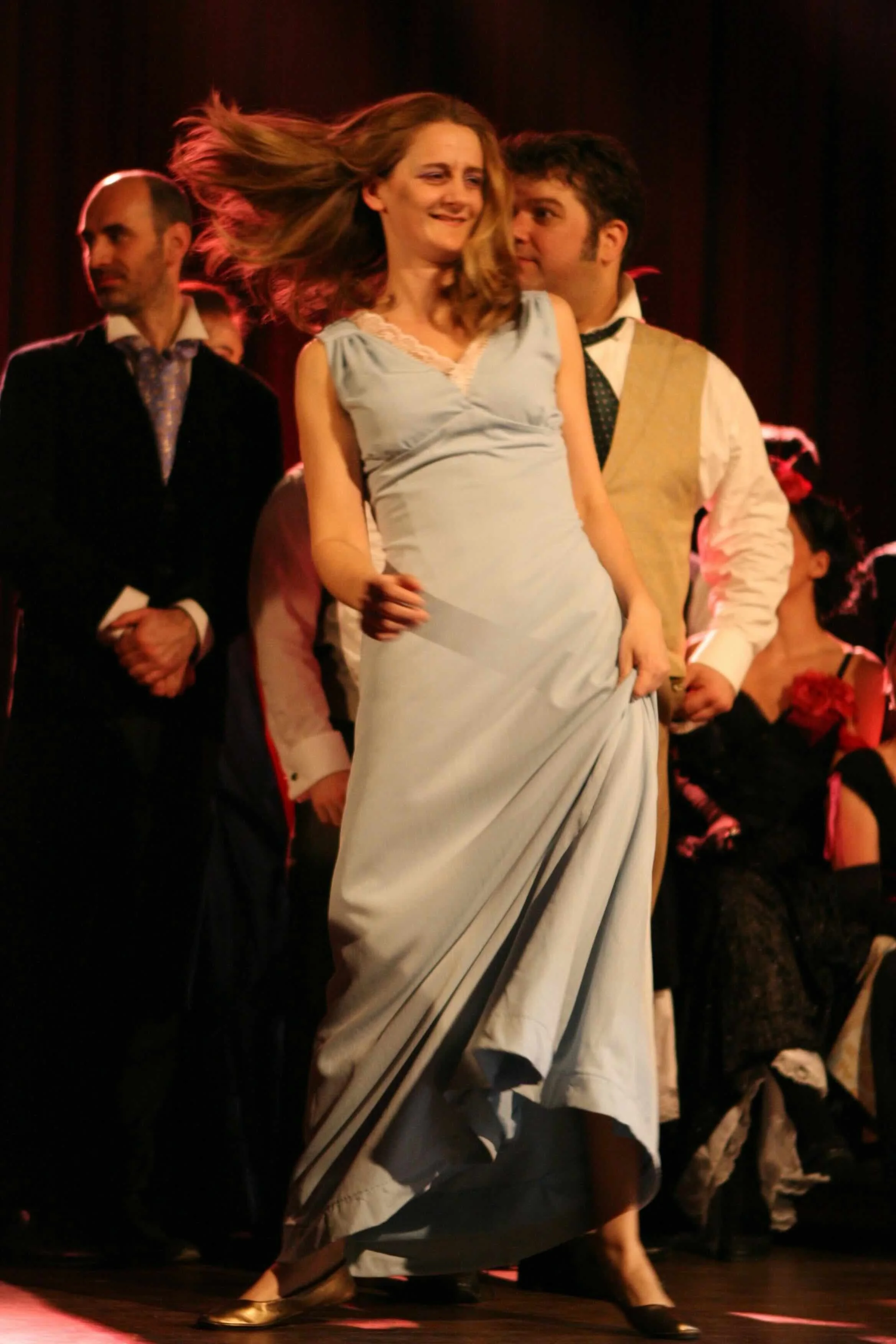 Sophie De Cruz en robe bleue dans une danse de scène, entourée d'autres personnes, sur scène avec lumière chaude.