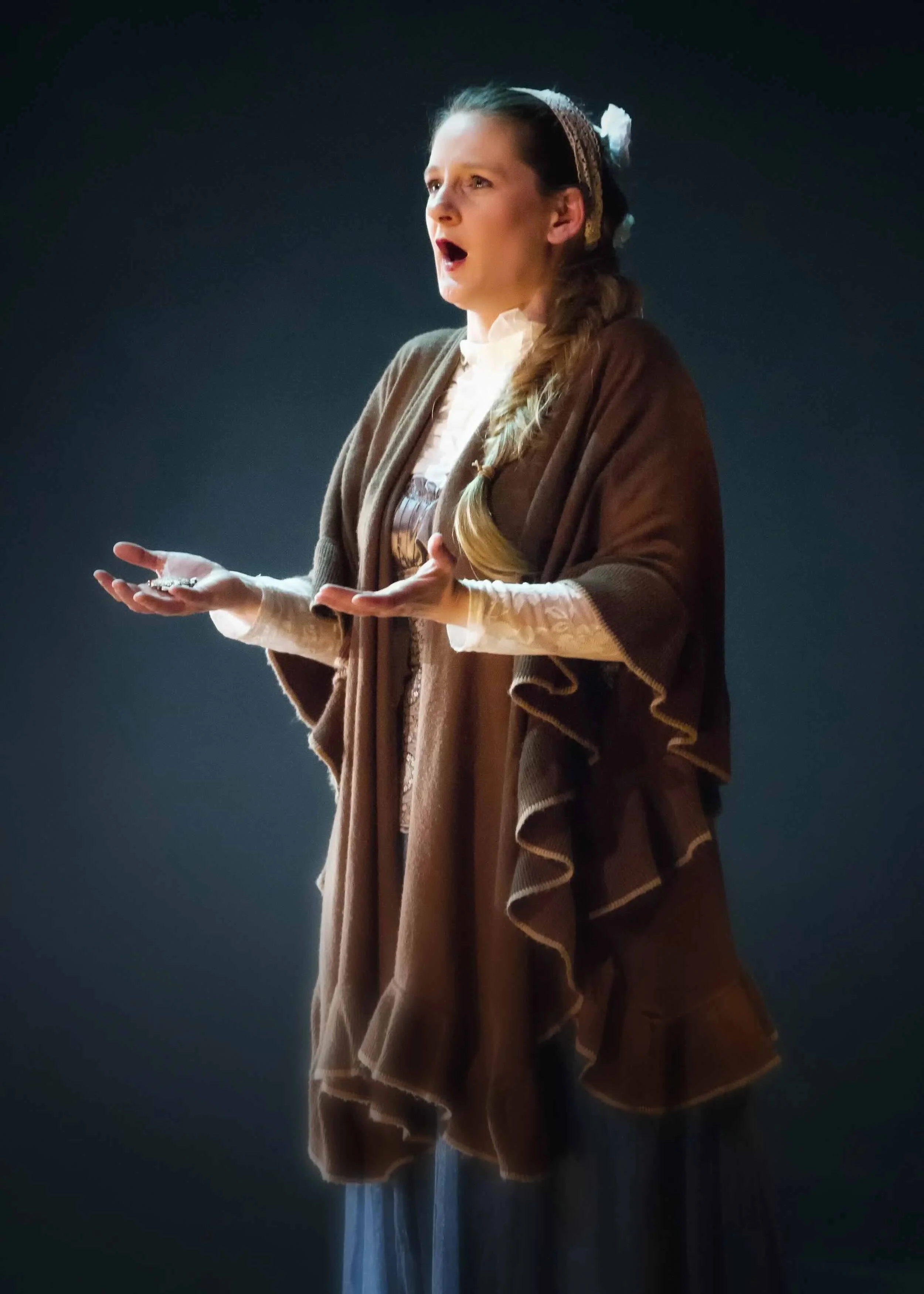 Sophie De Cruz en costume ancien avec des cheveux tressés et un bandeau, en pleine performance théâtrale ou chant, avec un fond sombre.