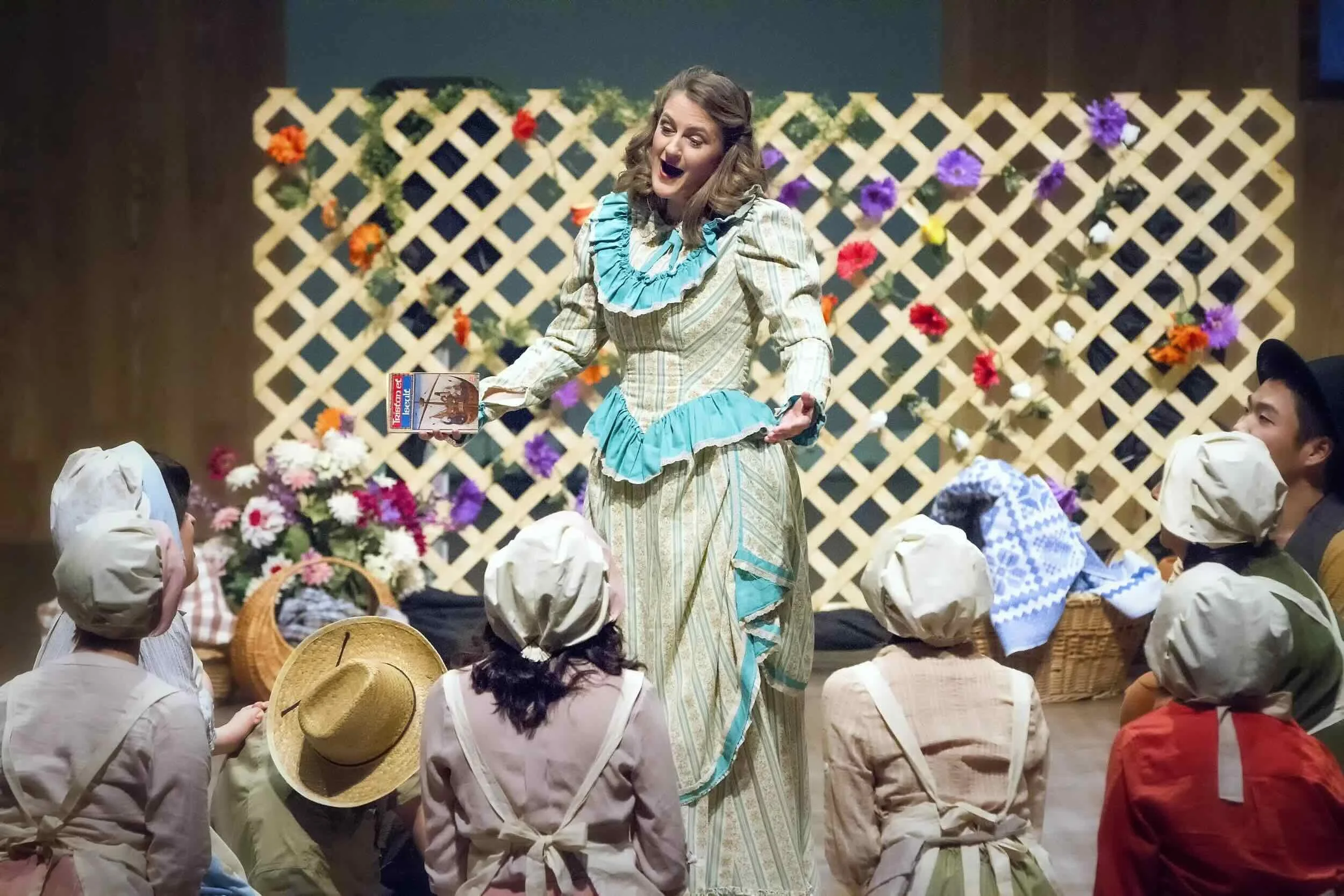 Sophie De Cruz en costume vintage avec un collier bleu, debout devant un groupe de personnes en vêtements anciens, dans un décor avec une clôture en bois décorée avec des fleurs colorées.