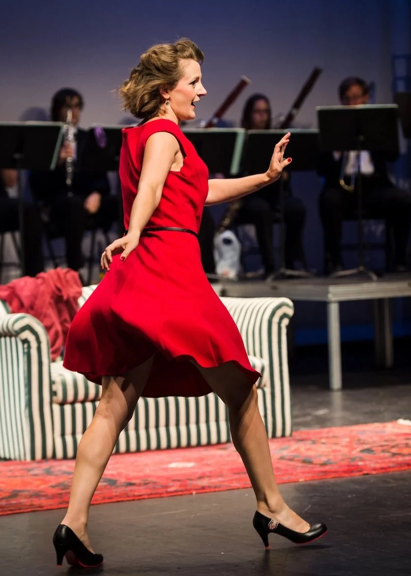 Sophie De Cruz en robe rouge danse ou joue sur scène, avec un orchestre en arrière-plan.