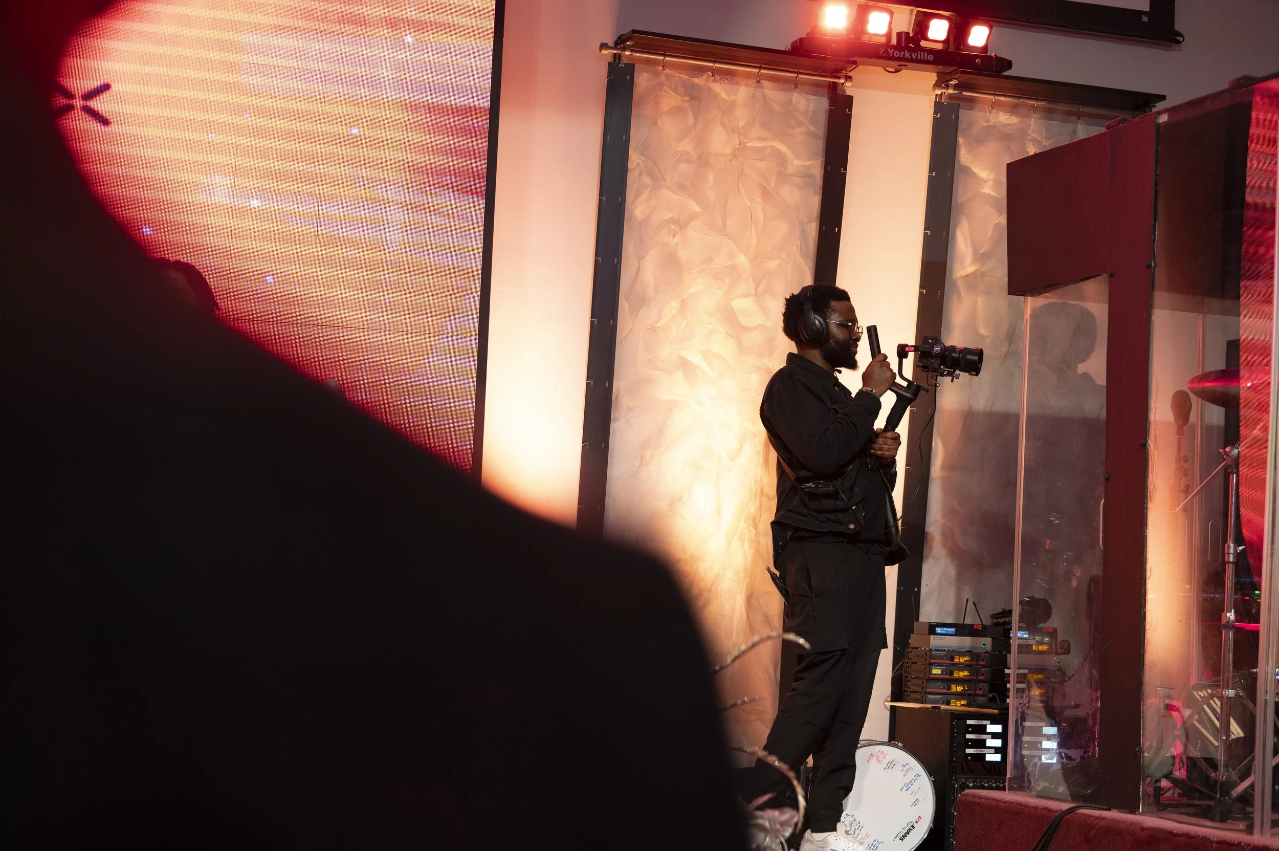 A videographer holding a camera and microphone on a stage behind a glass partition, with colorful lighting and large screens in the background.