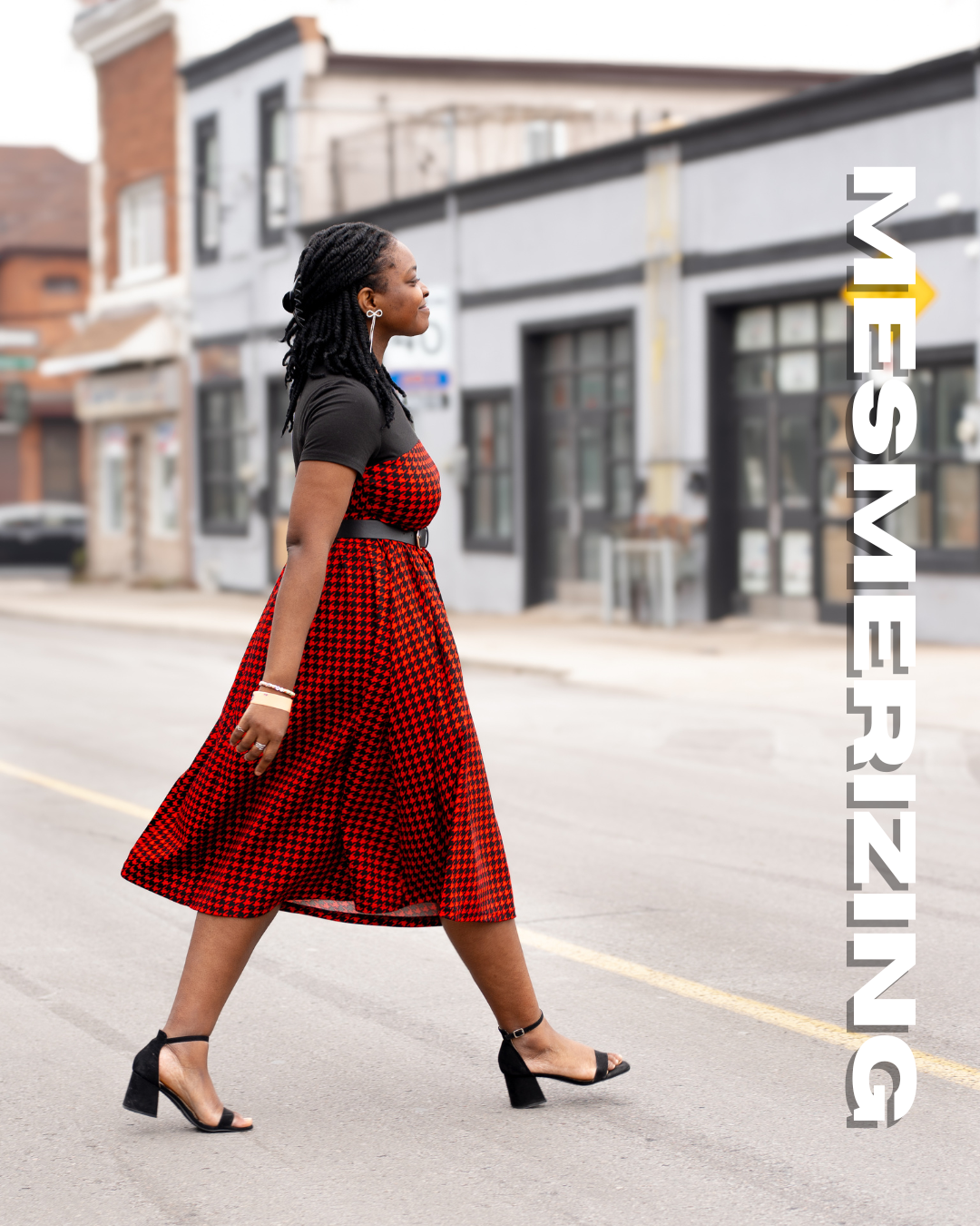 A woman walking across a street in an urban area, wearing a red and black dress, black top, black sandals with a block heel, and earrings. The background features storefronts and buildings.