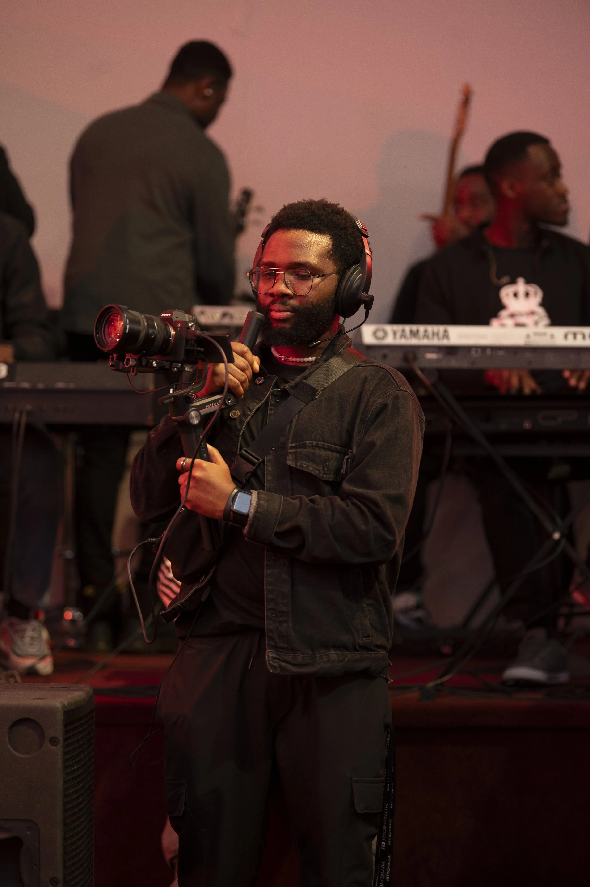 A man in a black jacket with headphones and a camera, holding a microphone, standing near a stage with musicians in the background.