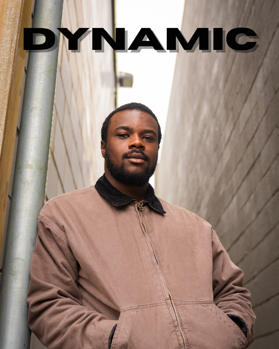 A man with dark skin, a beard, and short hair stands between two tall concrete walls, wearing a tan jacket with his hands in the pockets. The word "DYNAMIC" appears in large black letters at the top of the image.