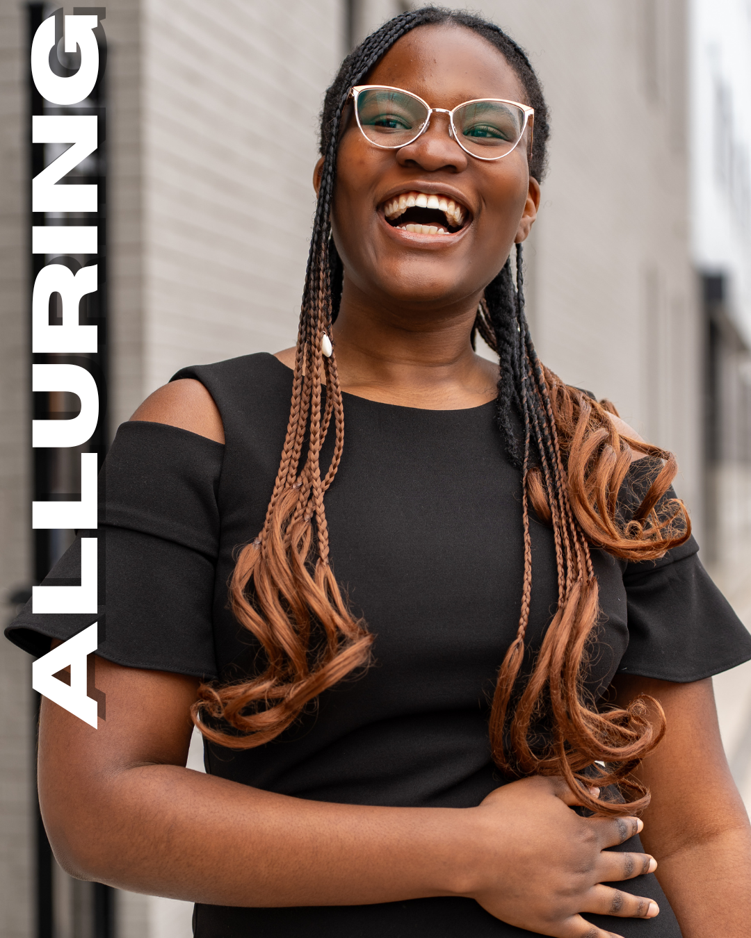 A woman with glasses, black and brown braided hair, smiling and wearing a black dress, standing outdoors near a brick building.