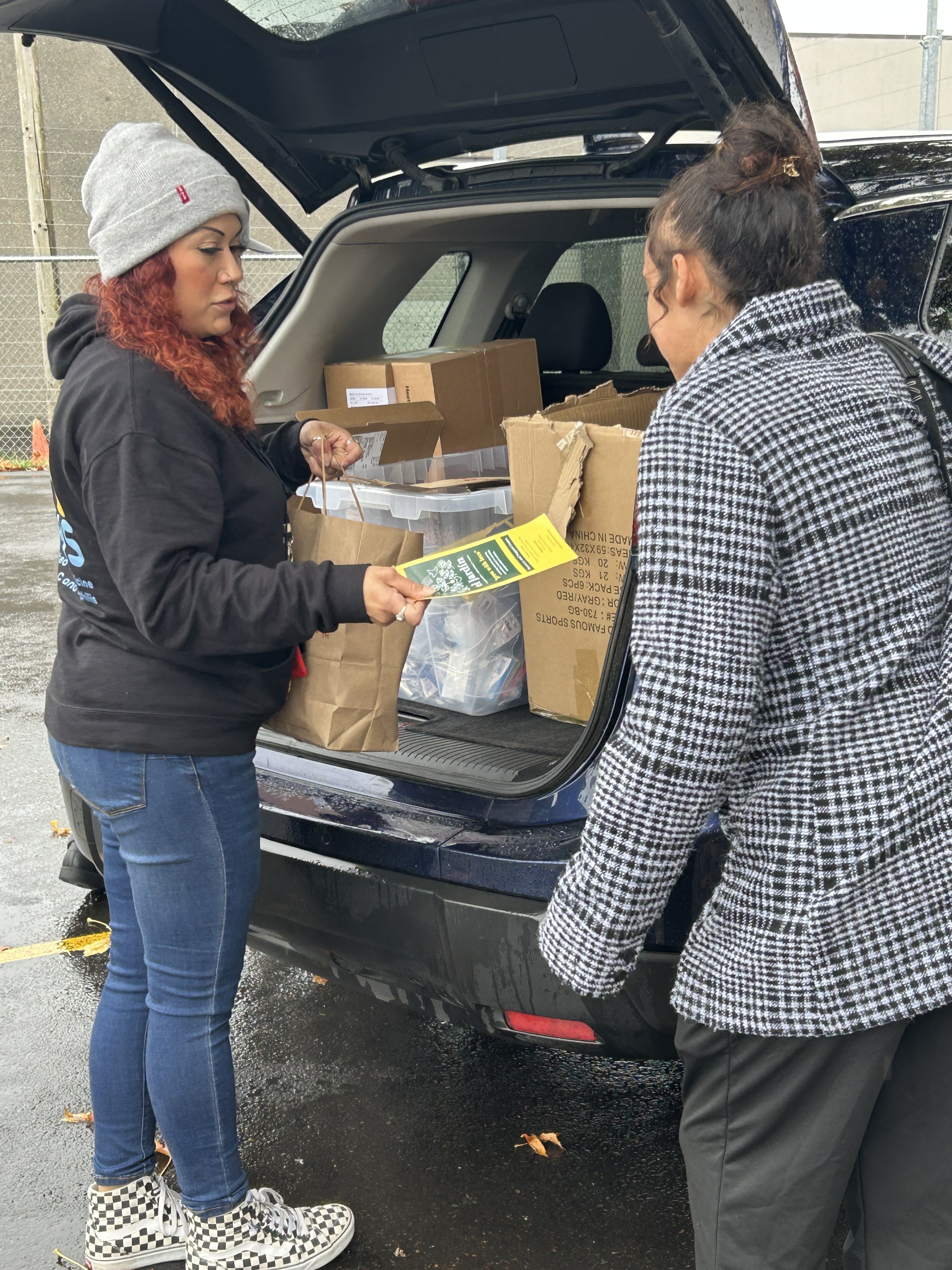 Two women stand at the open tailgate of a van full of harm reduction supplies. One woman stands with a bag of supplies and a flyer about El Jardin. She is speaking to the other woman about reccovery services, how to access them, and how to stay safe.