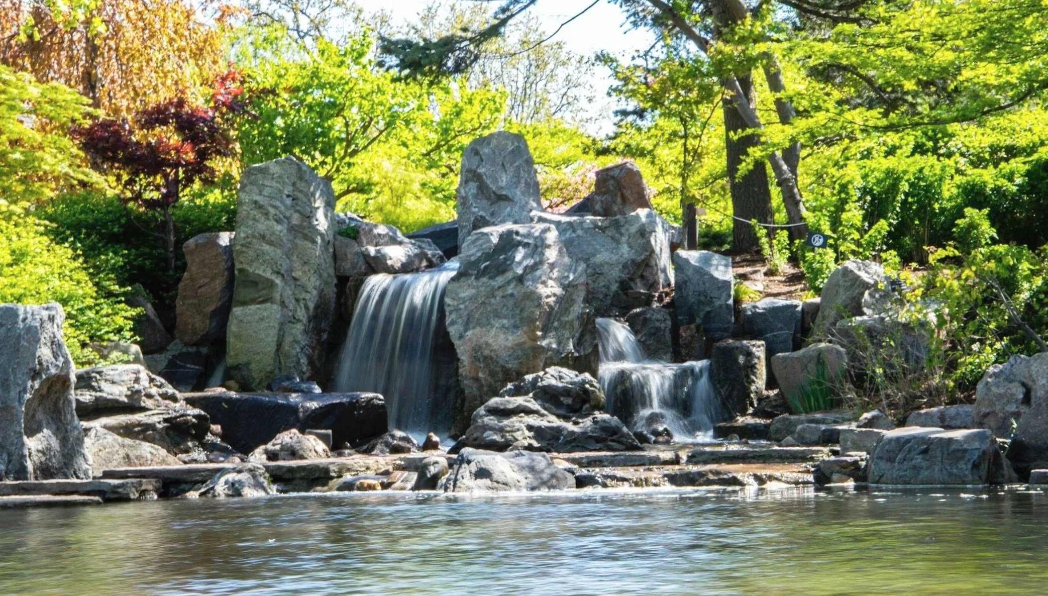 A small waterfall flowing over rocks into a pond surrounded by green trees and bushes.
