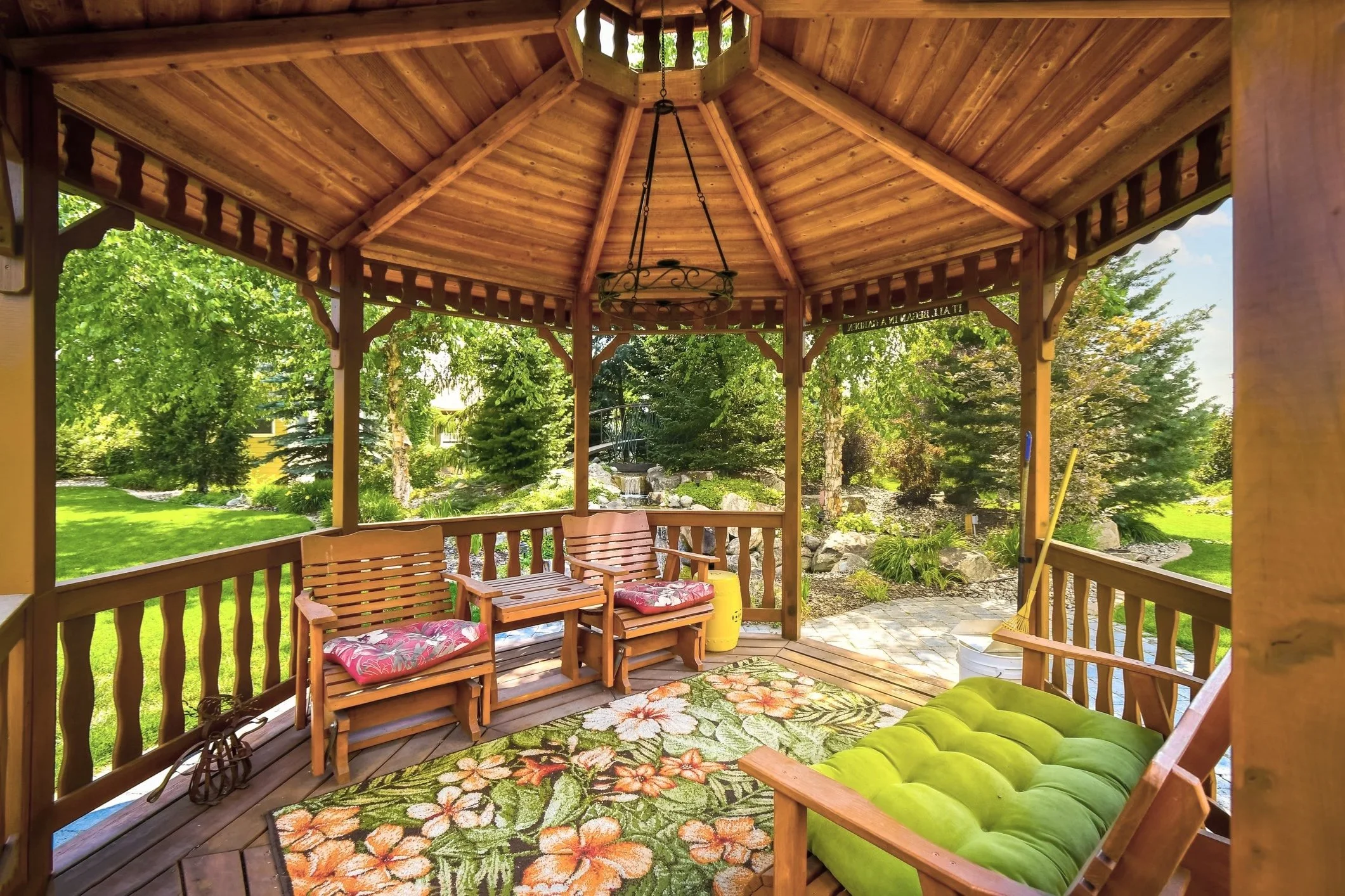 A wooden gazebo with a floral rug, two chairs with pink cushions, a green cushioned bench, and a bucket with cleaning tools, overlooking a garden with lush green trees and shrubs.