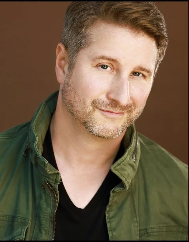 Man with short, light brown hair and a beard wearing a green jacket and black shirt, smiling towards the camera.
