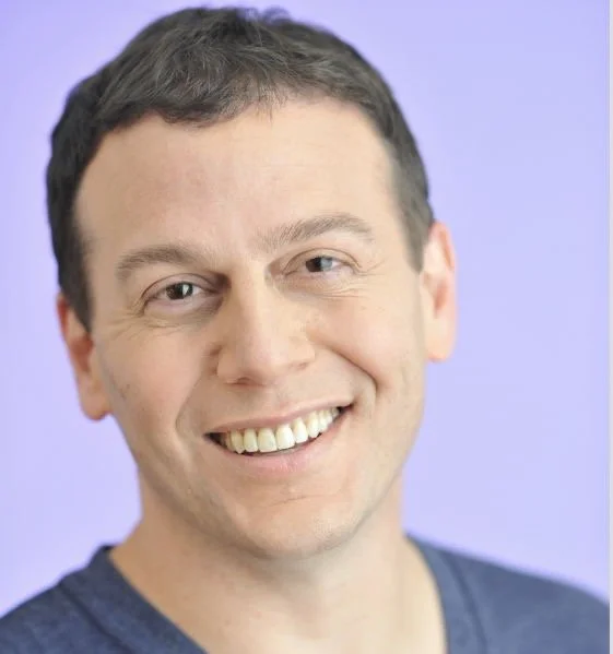 A smiling man with short dark hair, wearing a dark blue shirt, in front of a purple background.