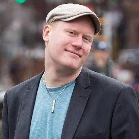 A young man wearing a gray cap, blue shirt, black blazer, and a silver necklace, smiling outdoors.