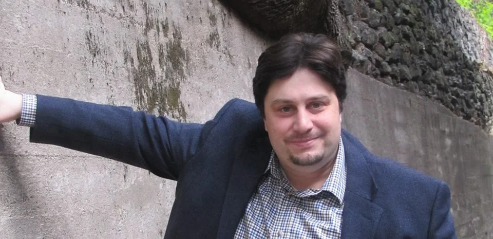 A man with dark hair, wearing a checked shirt and a navy blazer, smiling while standing outdoors against a concrete wall and rocky terrain.