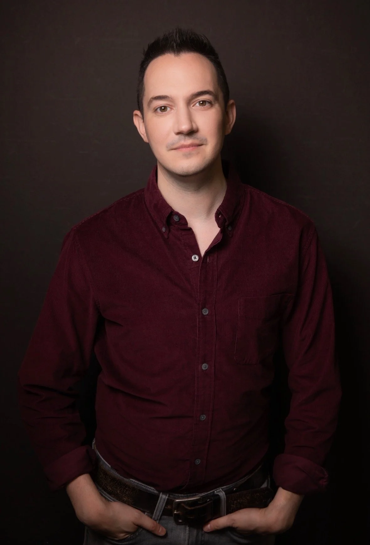Portrait of a young man with dark hair wearing a maroon button-up shirt against a dark background.