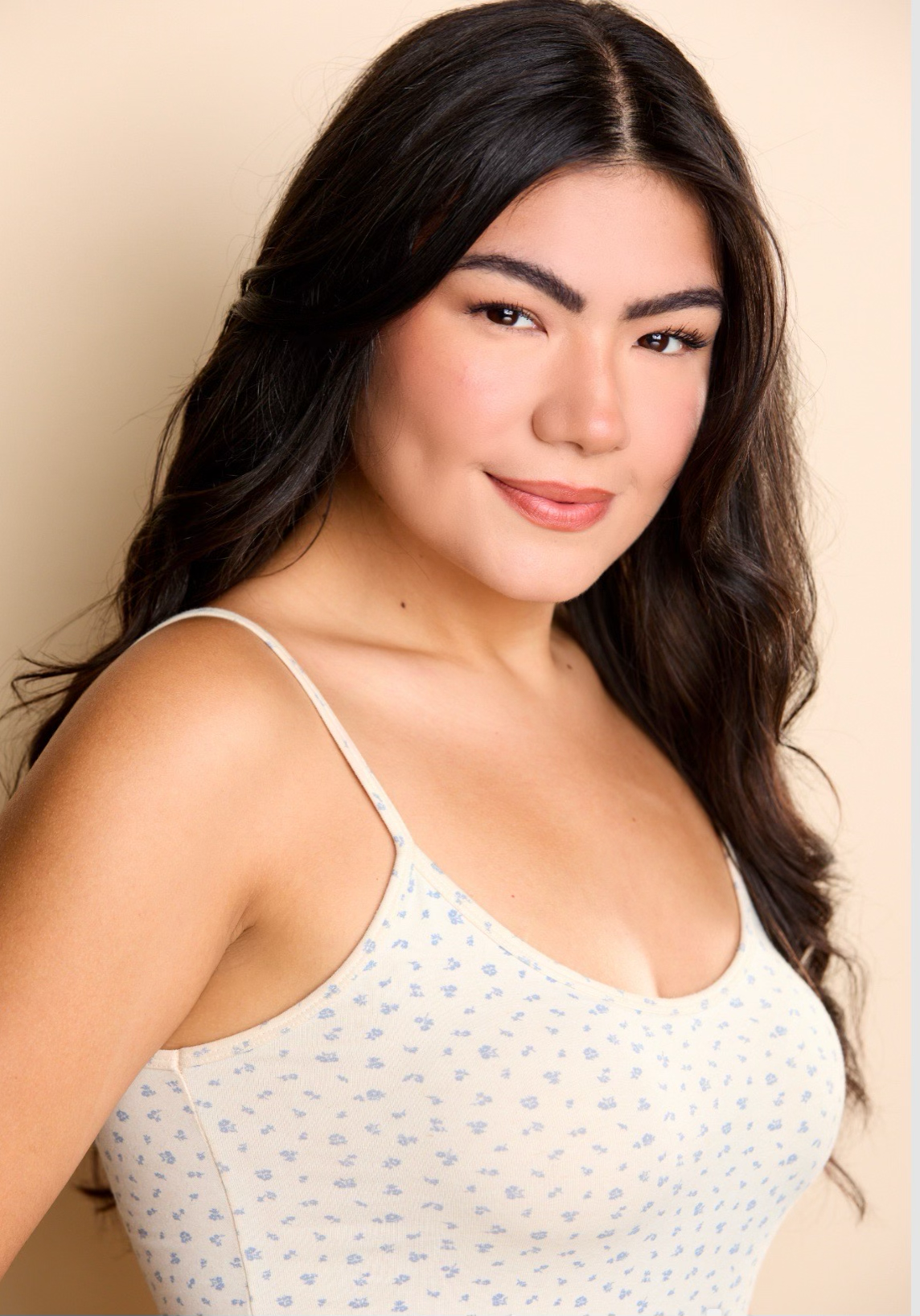 Woman with long dark hair wearing a light floral tank top, smiling at the camera against a beige background.
