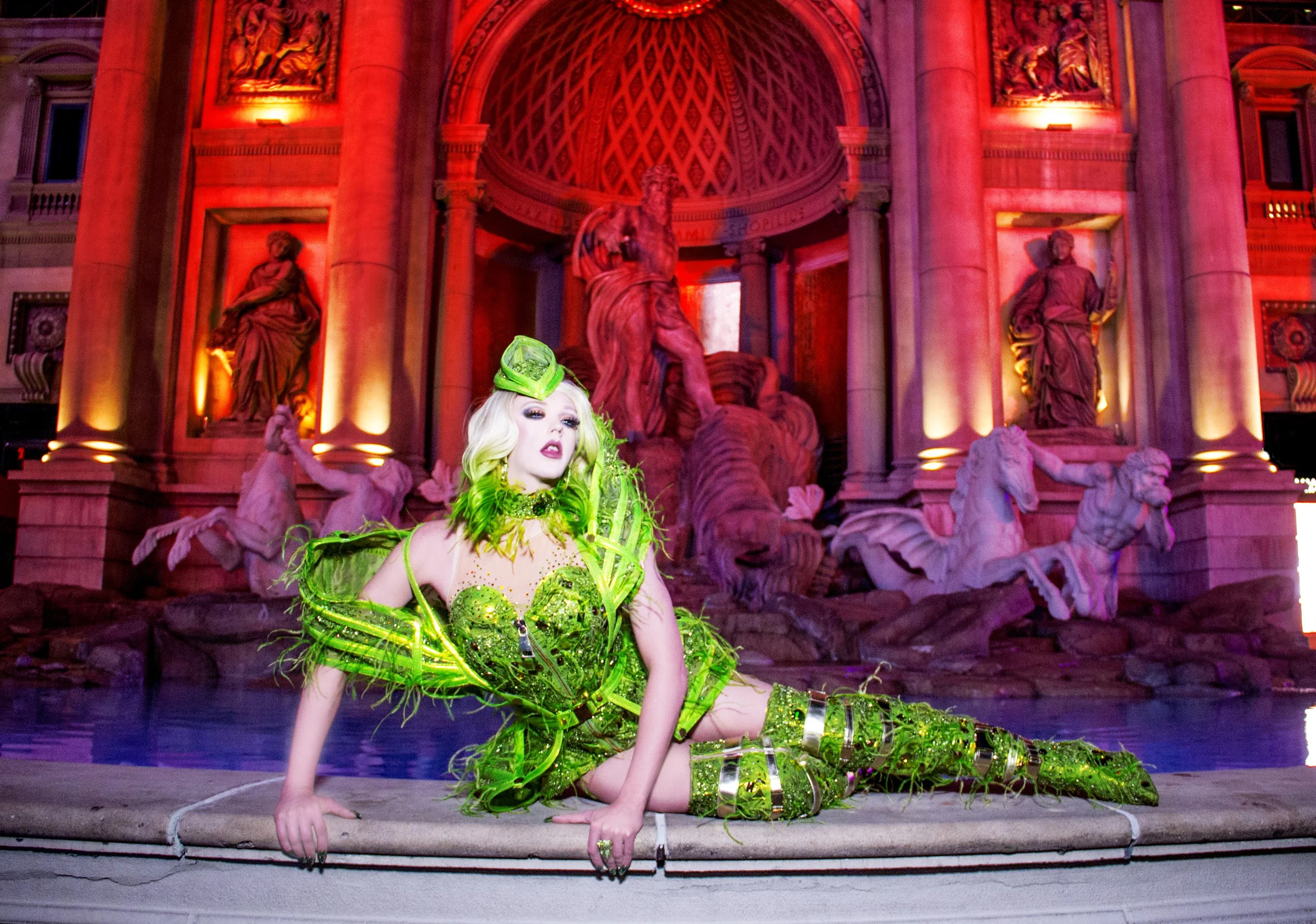 A person dressed in bright green outfit with futuristic accessories, posing on stage in front of a large Baroque-style marble fountain with statues, illuminated with purple and orange lighting.
