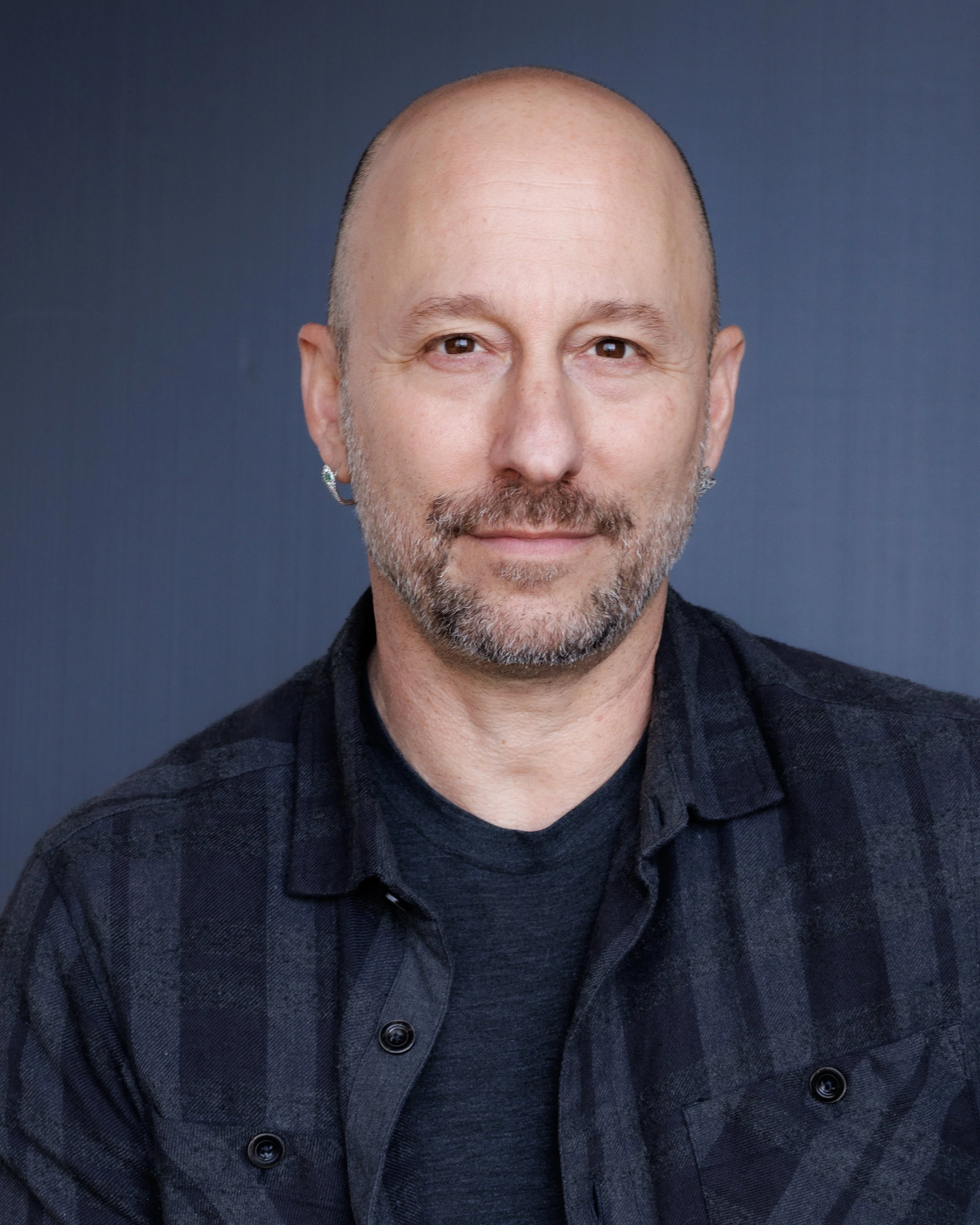 Close-up portrait of a middle-aged man with a bald head, brown eyes, and a short beard, wearing a black and gray plaid shirt against a dark blue background.
