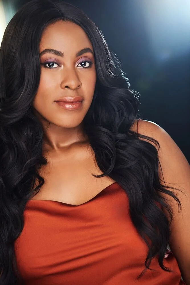 Portrait of a woman with long black hair wearing an orange sleeveless top, with a soft focus background and light flare.