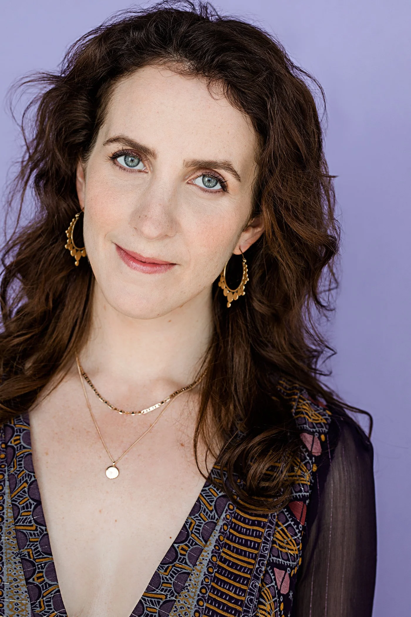 Close-up of a woman with long, curly brown hair, blue eyes, wearing gold jewelry, and a patterned dark-colored top, against a purple background.