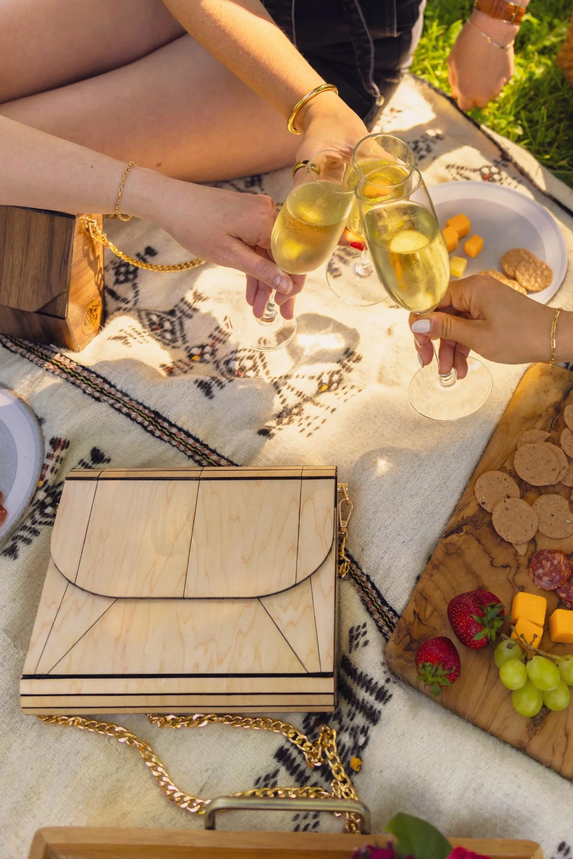 People clinking glasses of sparkling wine during a picnic on a blanket with various snacks, cheese, strawberries, and grapes.