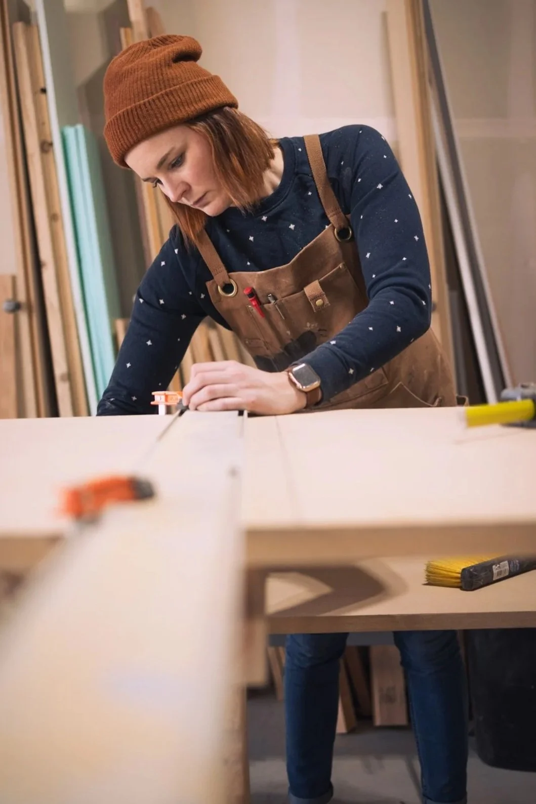 A woman wearing a brown beanie and overalls is working on a woodworking project in a workshop. She is focused, measuring or marking a piece of wood on a workbench.