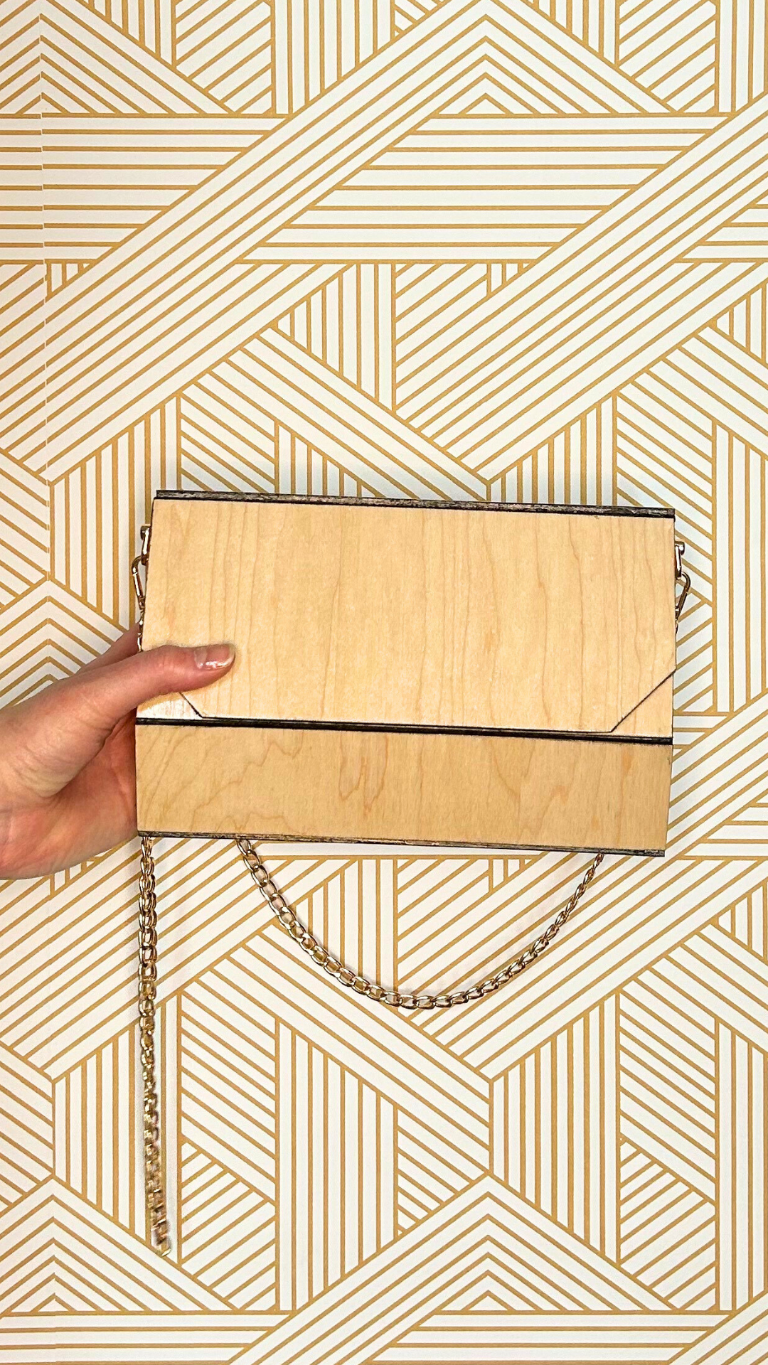 A wooden clutch purse with a chain strap resting on patterned gold and white geometric wallpaper. A hand with a light nail polish holds the clutch on the left side.