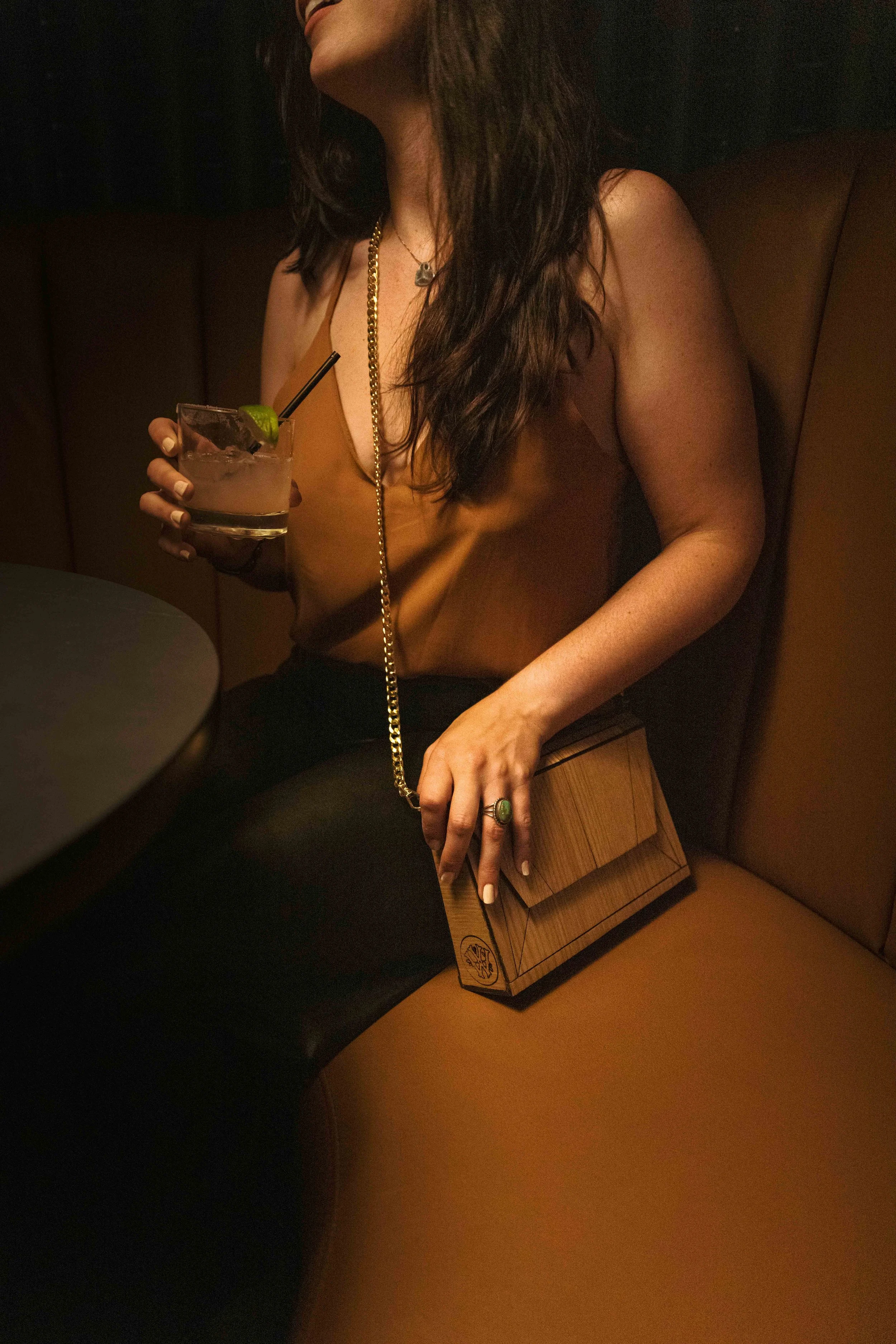 A woman sitting in a dimly lit bar or restaurant, holding a cocktail with a lime garnish. She is wearing a sleeveless top, a necklace, and a ring, and has long, wavy hair. There is a wooden box or clutch on her lap.