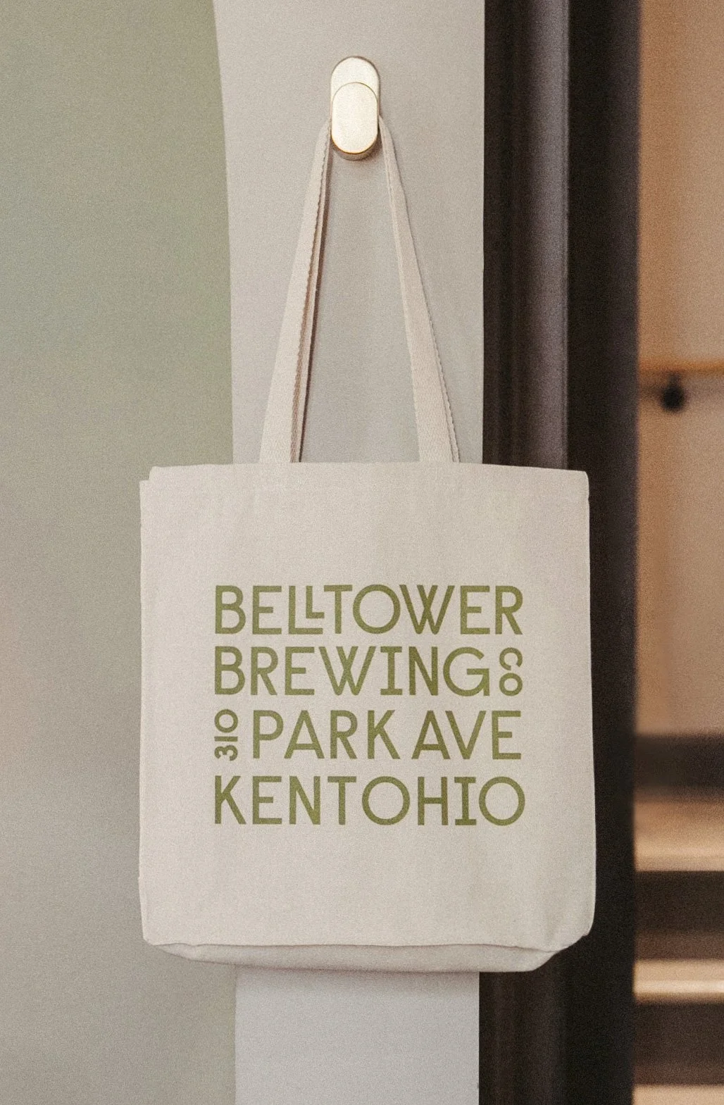 A beige tote bag hanging from a hook on a beige wall. The bag has green text that reads "BELTOWER BREWING 310 PARK AVE KENT OHIO."