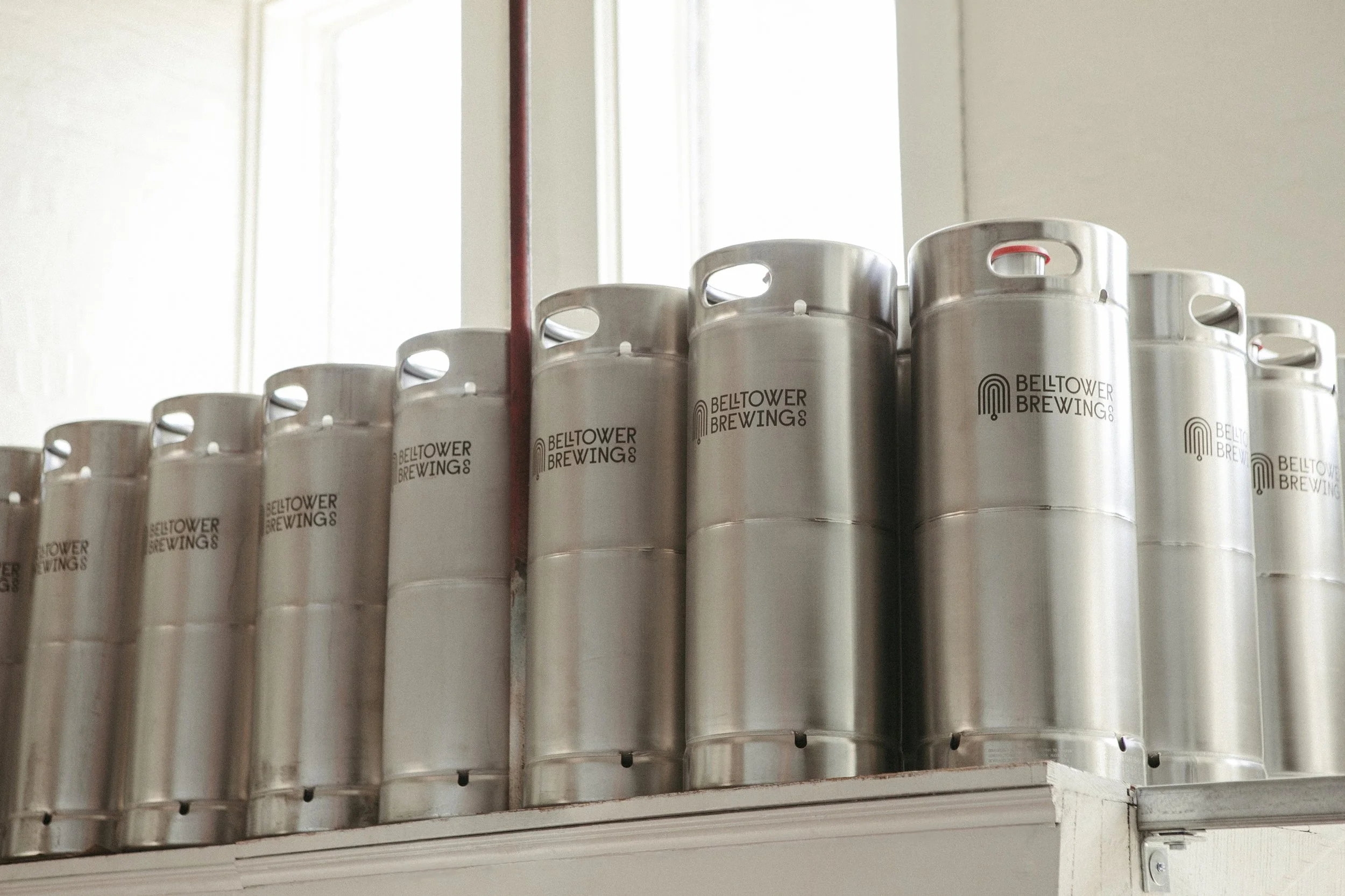 Stacked aluminum beer kegs labeled Belltower Brewing Co, stored on a shelf in a brewery.