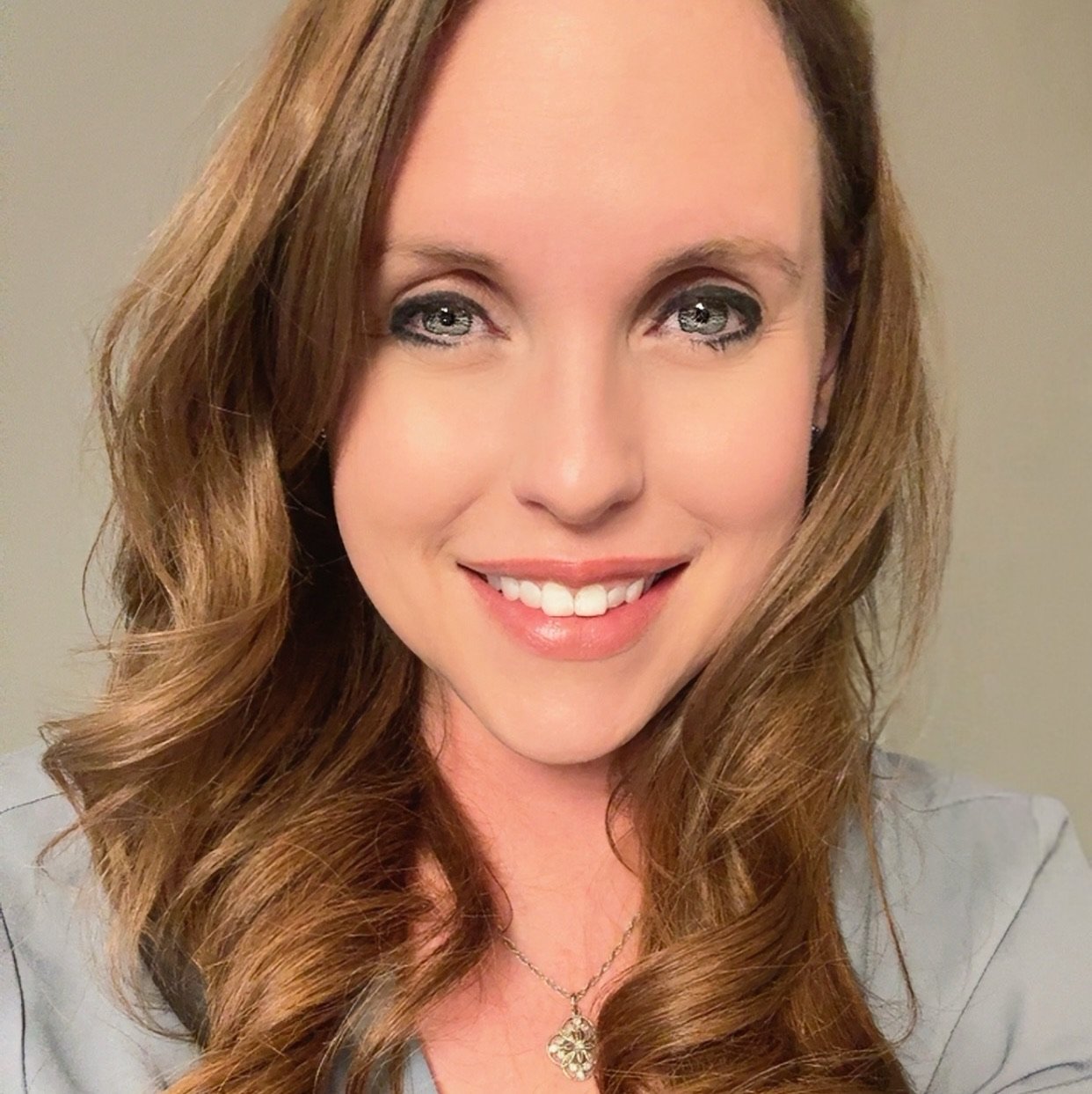 Close-up portrait of a smiling woman with curly, light brown hair and blue eyes, wearing a black top, a pearl necklace, and makeup.