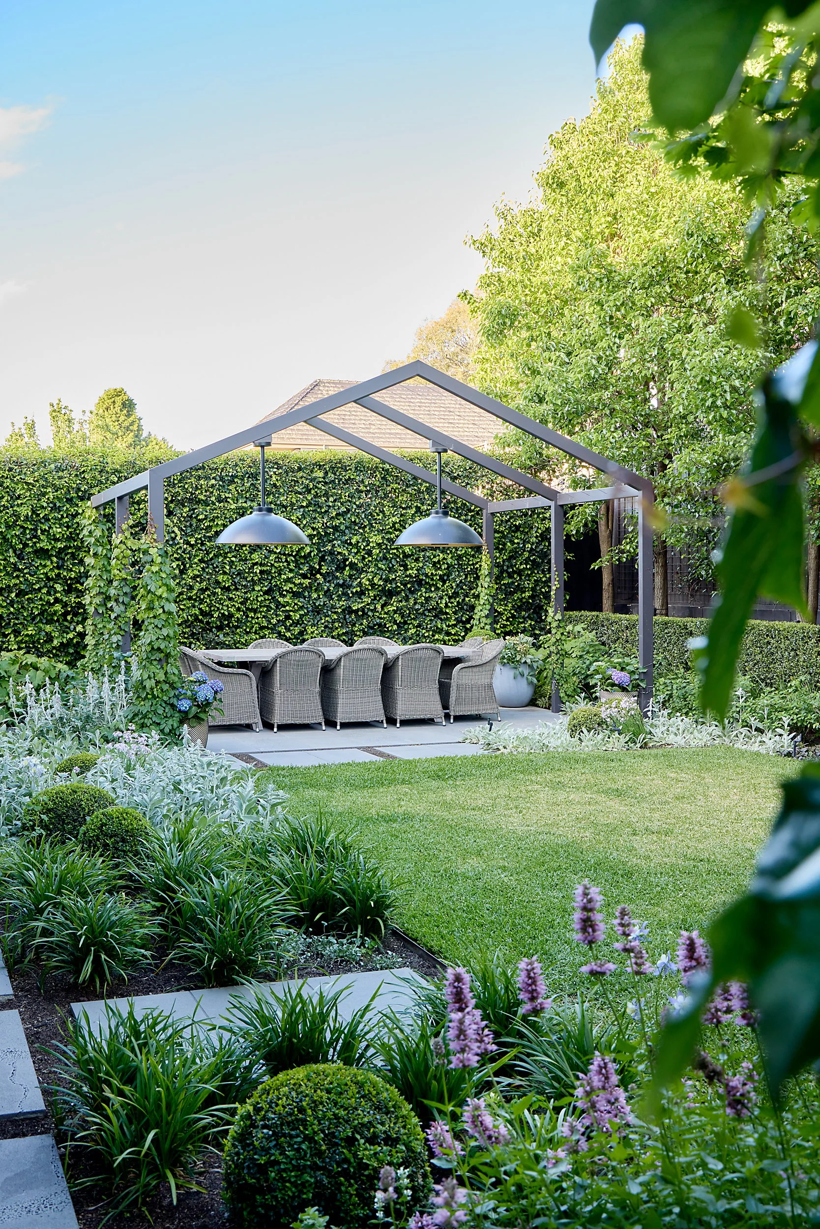 A backyard garden with a wooden pergola and hanging lights, surrounded by lush green plants, blooming flowers, and a well-maintained lawn.