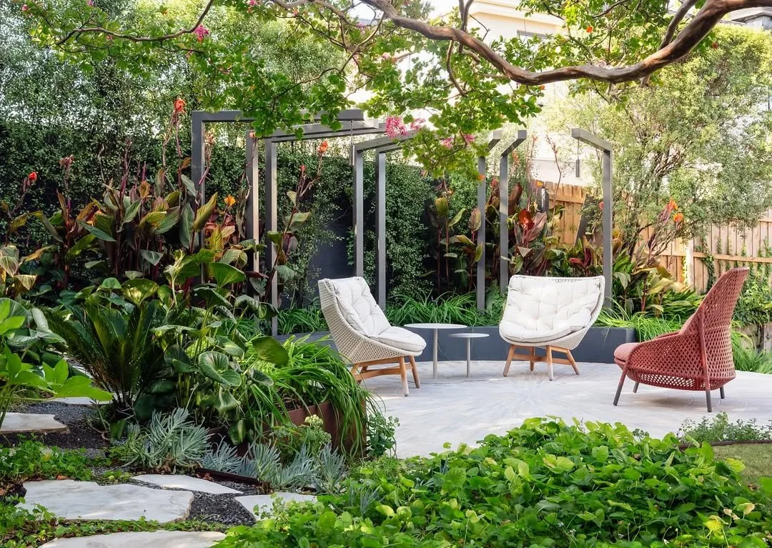 A backyard garden with lush green plants, trees, and flowering bushes. There are two modern white cushioned chairs and a red woven chair arranged around small side tables on a light-colored patio. Overhanging tree branches frame the scene, creating a cozy outdoor seating area.
