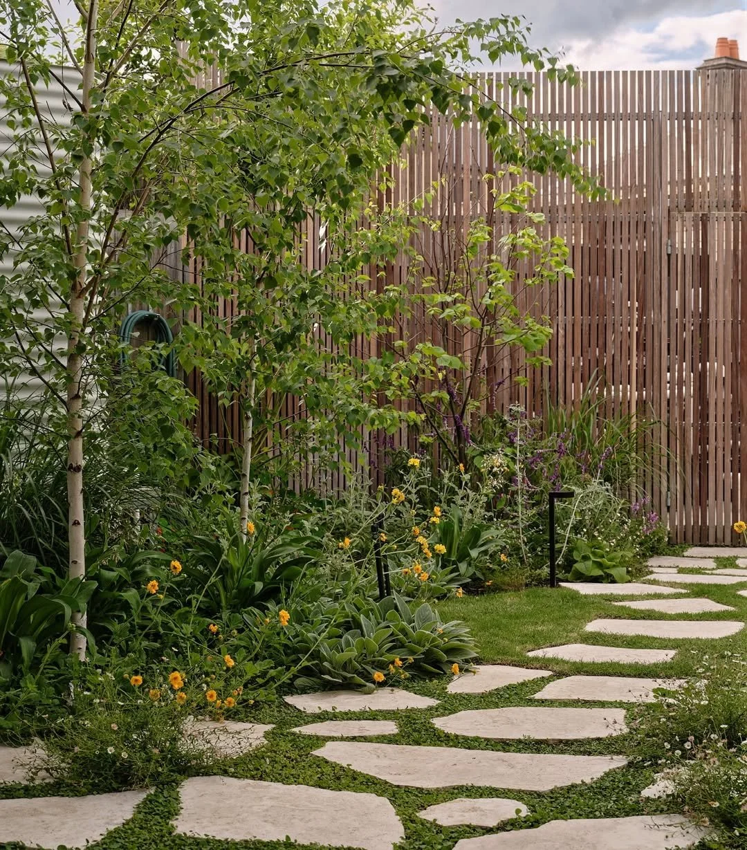 A garden pathway with stepping stones surrounded by lush green plants and trees, enclosed by a wooden fence.
