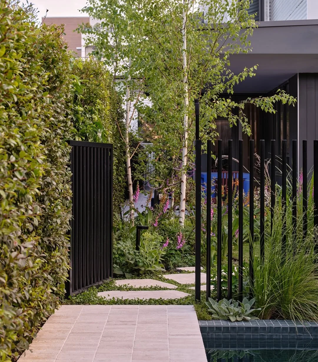 Modern backyard garden with stone pathway, lush greenery, trees, and black metal fencing, adjacent to a building with dark exterior walls.
