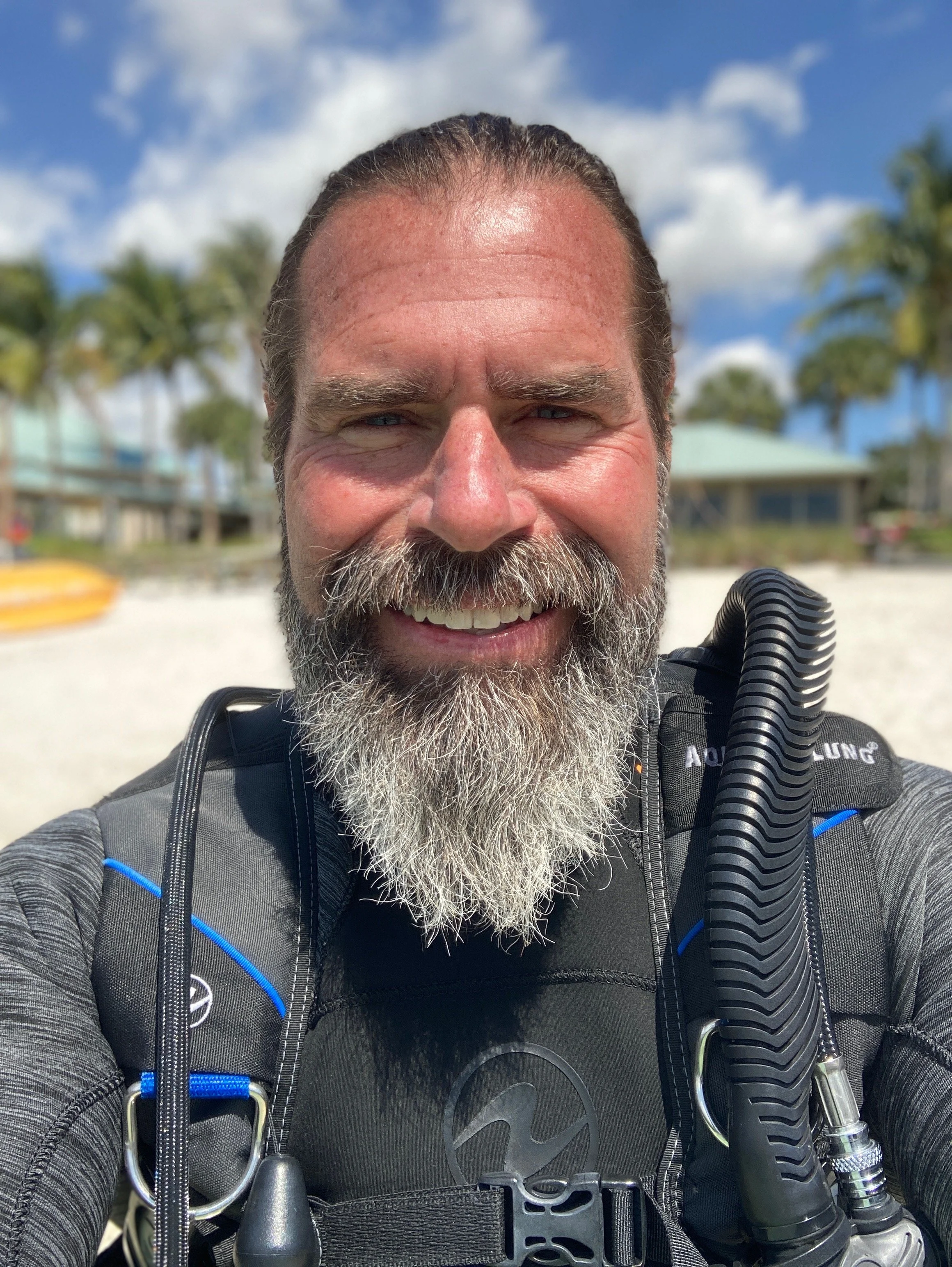 Man taking a selfie at the beach, smiling, with palm trees and a house in the background, wearing scuba gear.