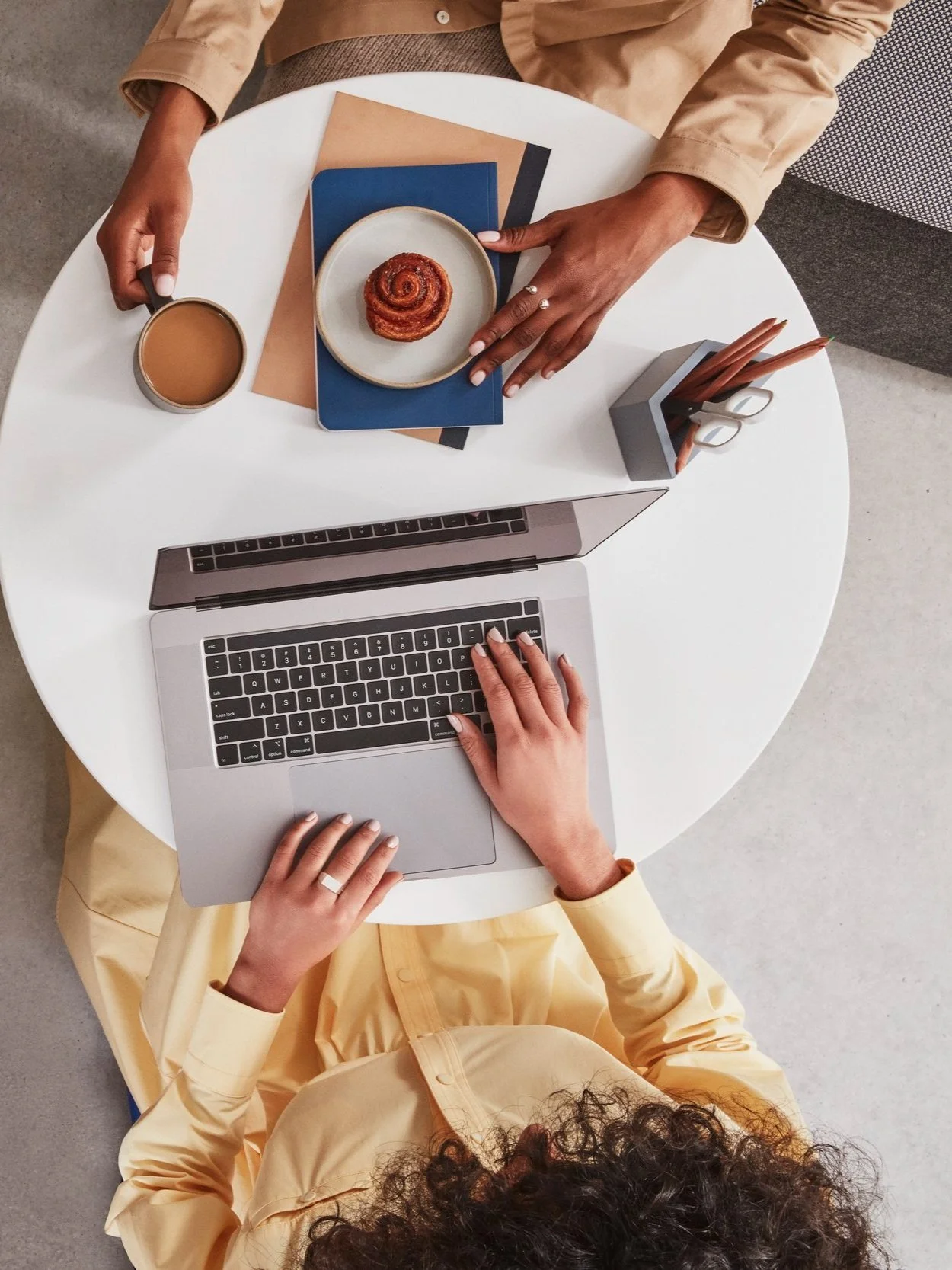 An image taken from above showing the hands of a Black lady and a White lady. The White lady is typing on her laptop and the Black lady has a cinammon bun on top of a book. At the side of them are a cup of coffee and a pencil pot.