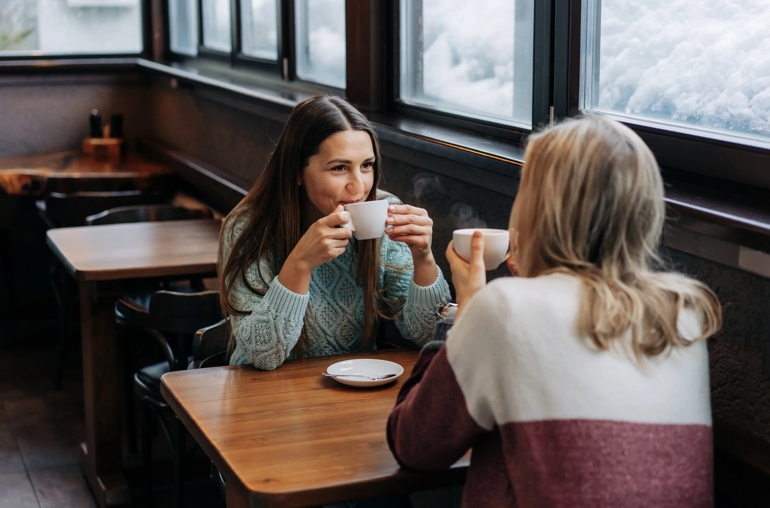 Image of a woman with dark eyes and dark, long hair wearing a light blue coloured cable knit jumper. She is sipping coffee and smiling sat opposite a friend.