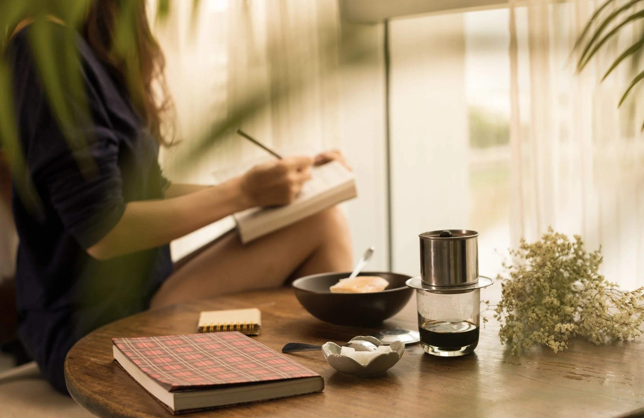 Image of a woman writing in a notebook in a cosy setting with just arms, torso and legs on show. On the table beside her is  a bowl of food and another book.