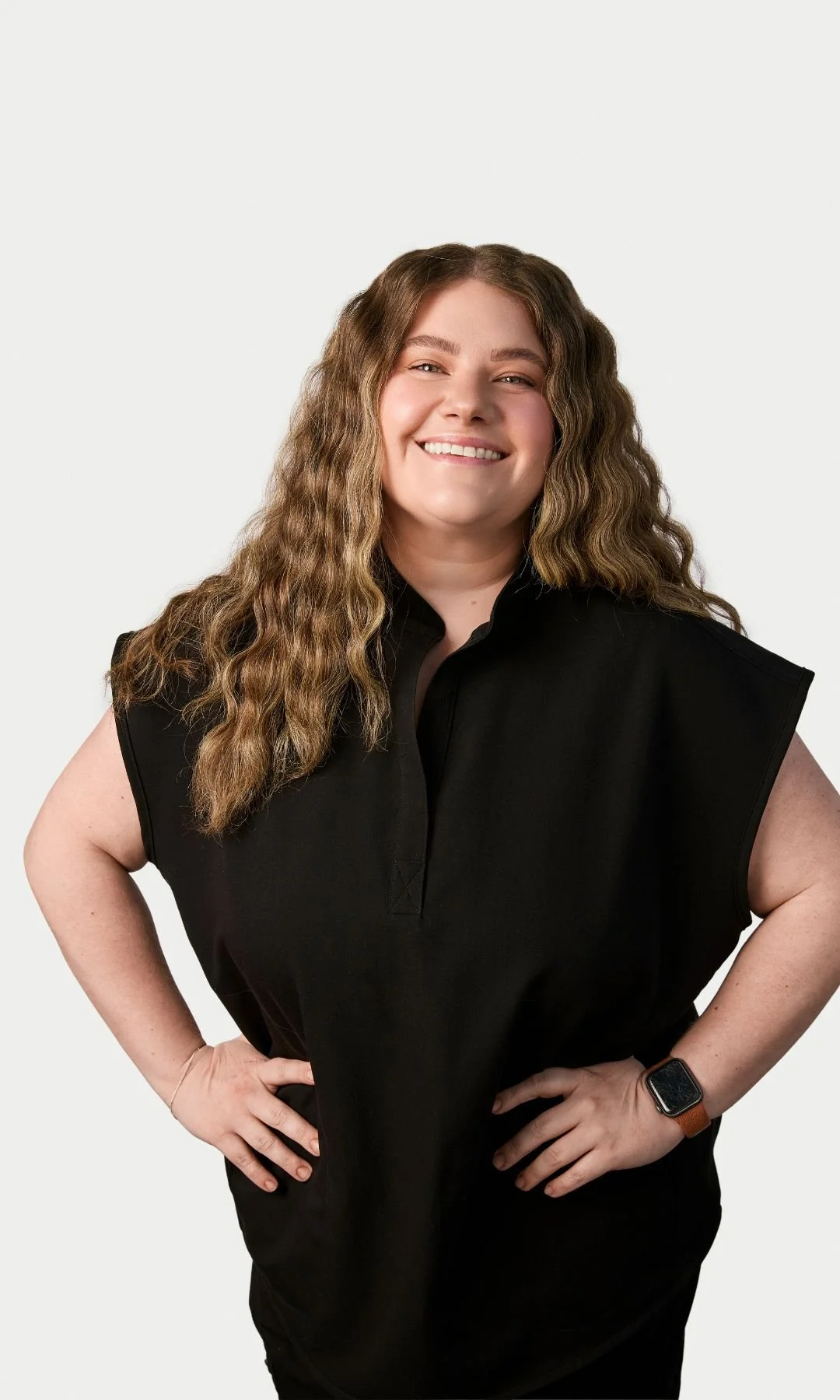 A woman with long wavy hair smiling, wearing a black sleeveless top and a smartwatch on her wrist, standing with hands on hips against a plain white background.