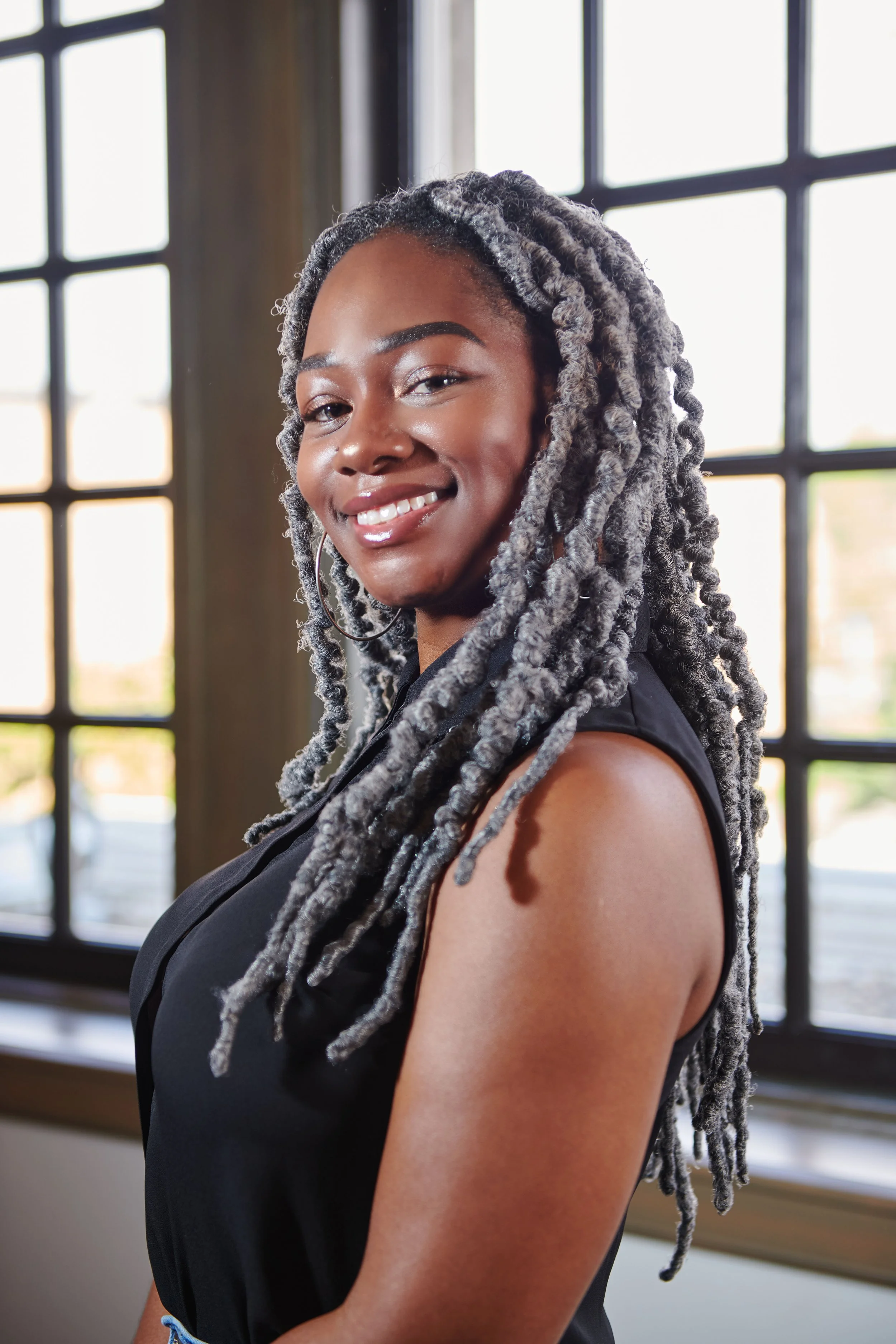 An environmental, corporate portrait of a pretty young woman with gray and black dreadlocks smiling indoors near a large window.
