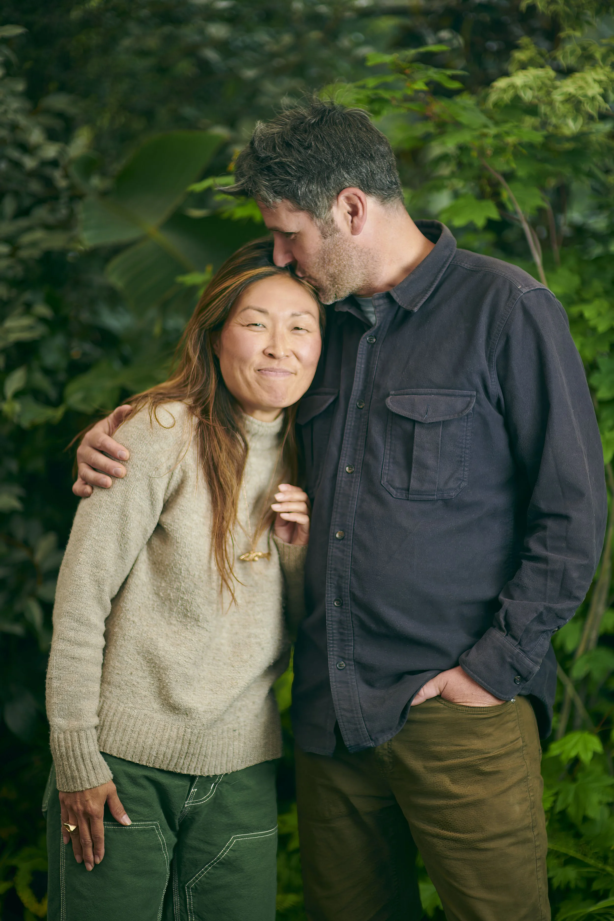 A man and woman sharing an affectionate moment in a lush green setting, with the man's forehead gently resting on the woman's head.