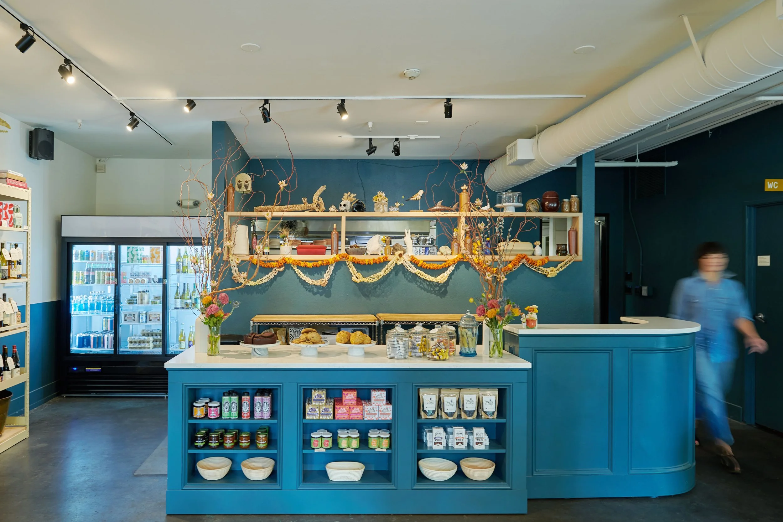 Interior of a small store or café decorated with orange and white garlands, featuring a central blue counter with jars, snacks, and flowers, and a back wall with shelves holding various items. A glass refrigerator with drinks is on the left, and a pe